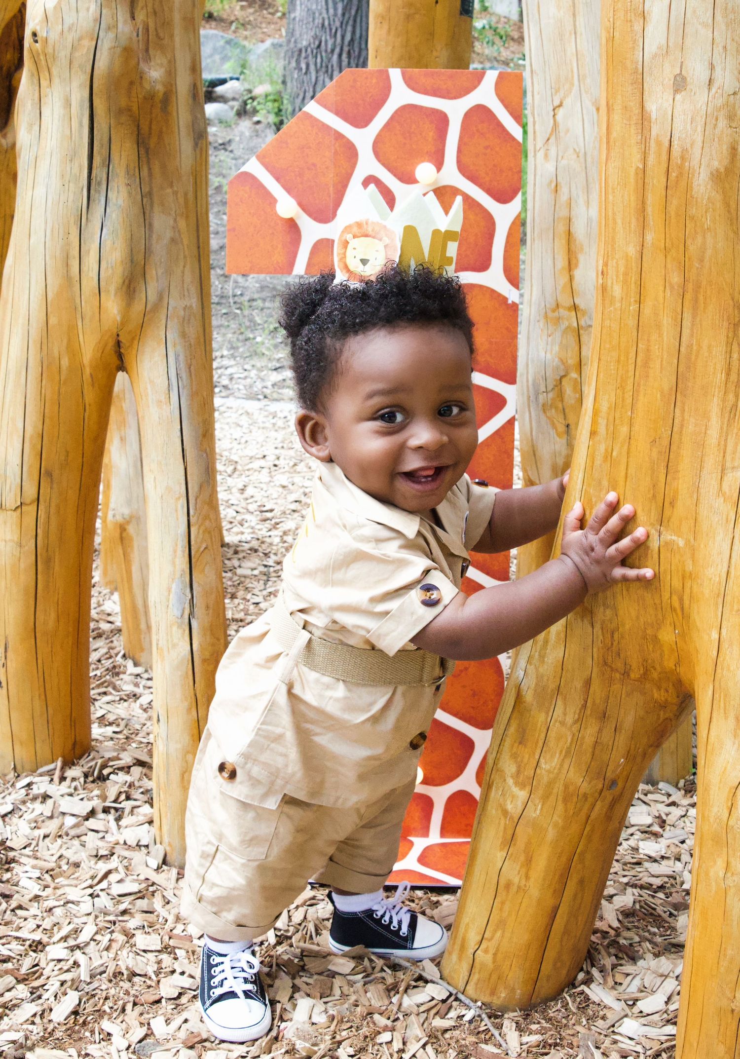 Smiling toddler in tan outfit holding wooden post at safari-themed playground with giraffe decoration.