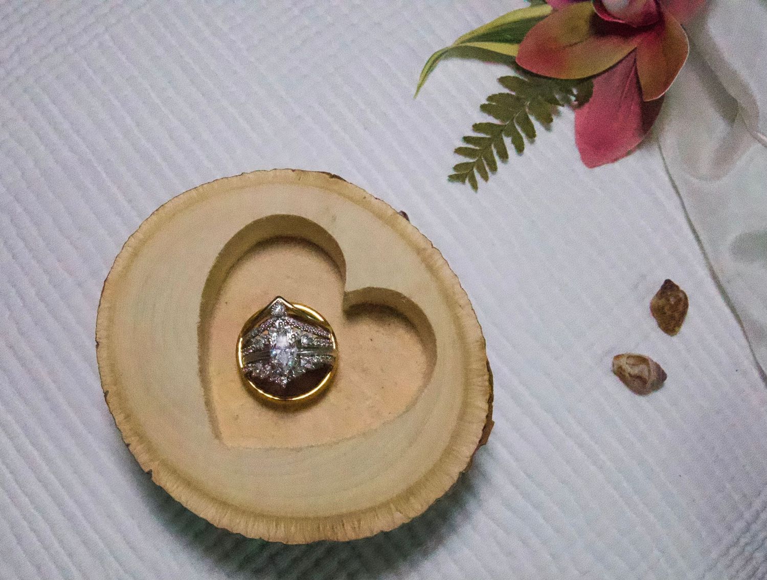 Gold diamond ring displayed in a rustic wooden heart-shaped ring dish, with pink flowers and fern leaves in background.