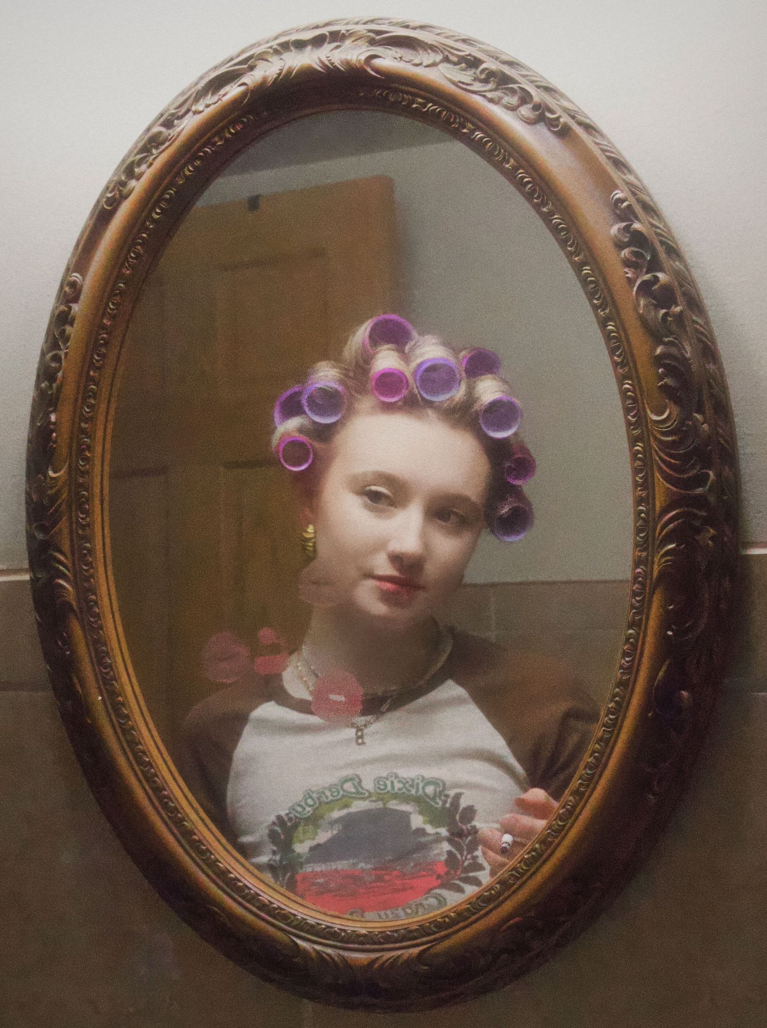 Young girl with purple hair rollers reflects in an ornate oval wooden framed mirror.