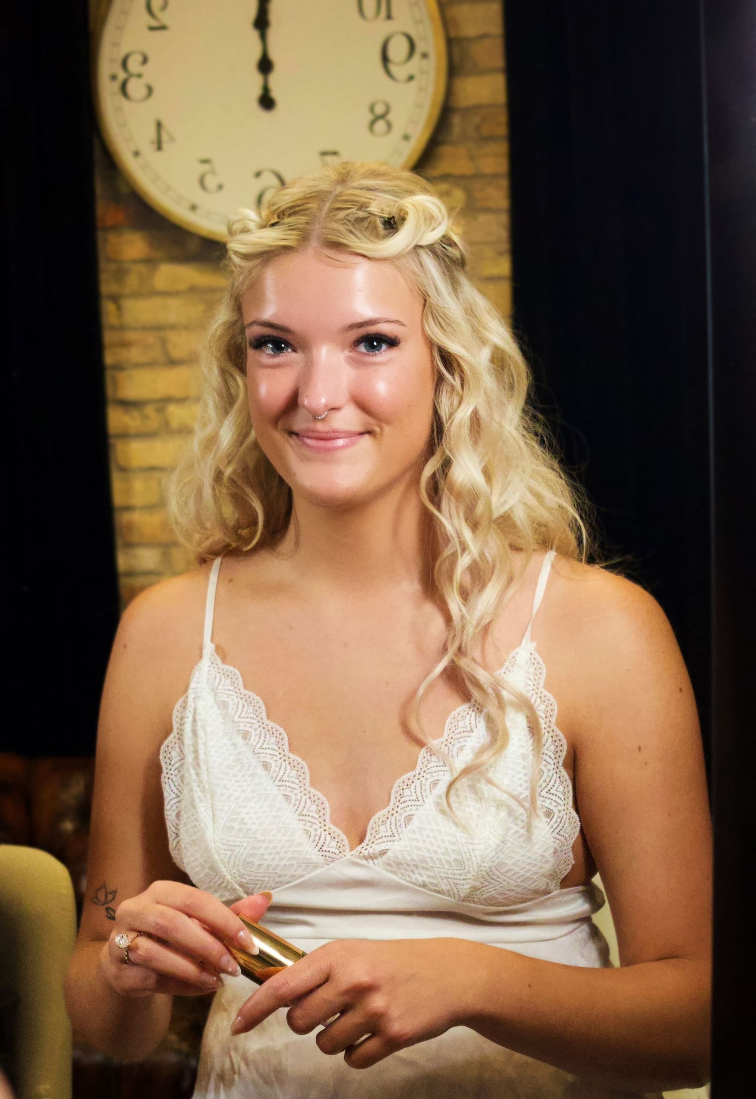 Smiling blonde bride in white lace dress holding lipstick, with vintage clock on brick wall background.