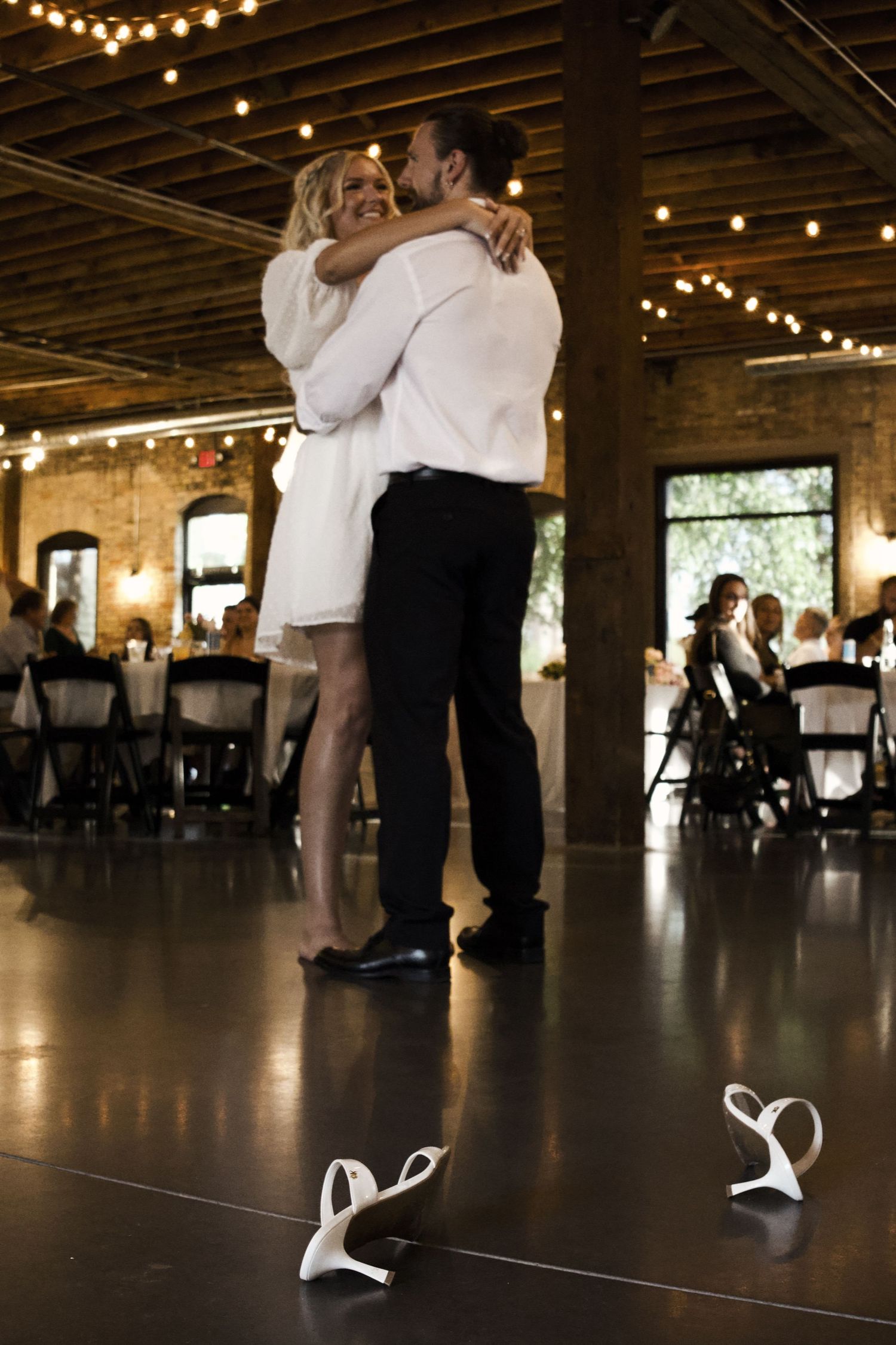 Bride and groom share first dance barefoot in elegant rustic wedding venue with string lights.