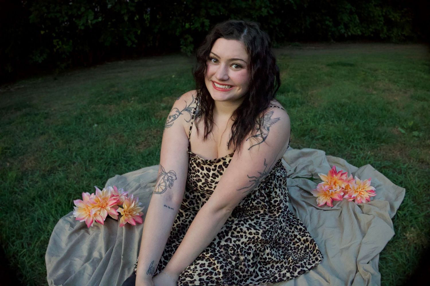 Woman in leopard print dress sits on grass with pink flowers, showing tattoos, smiling outdoors.