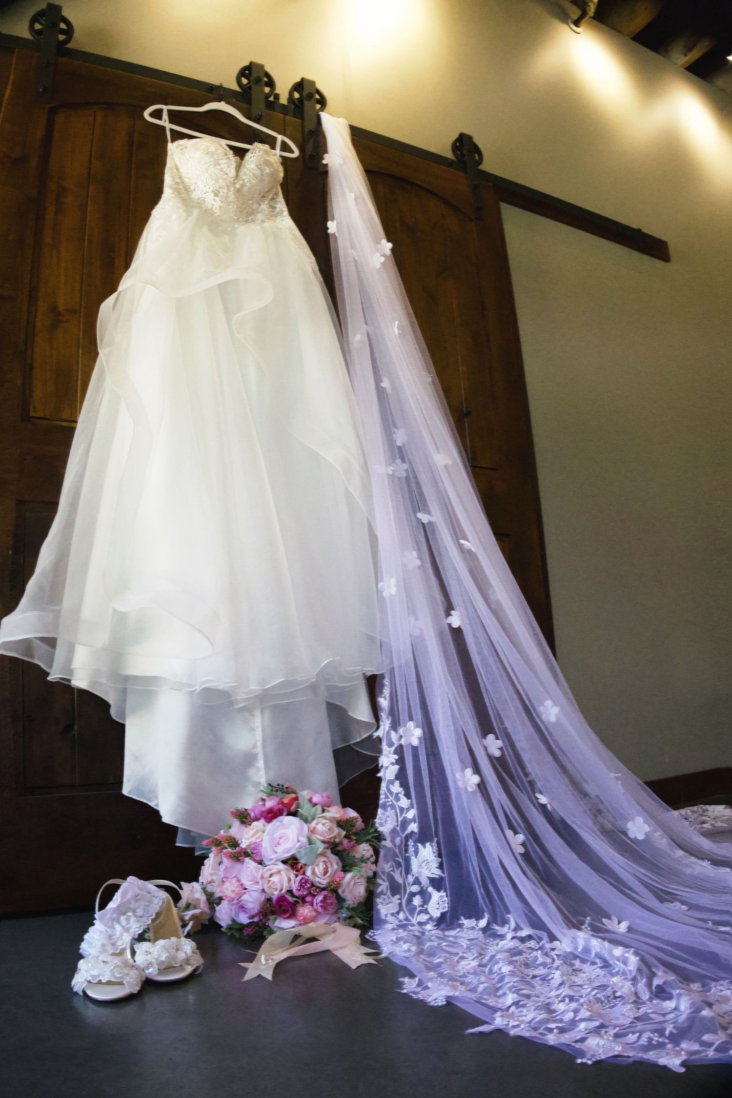 White wedding dress hanging on barn door with floral veil, bridal bouquet, and white heels on floor.