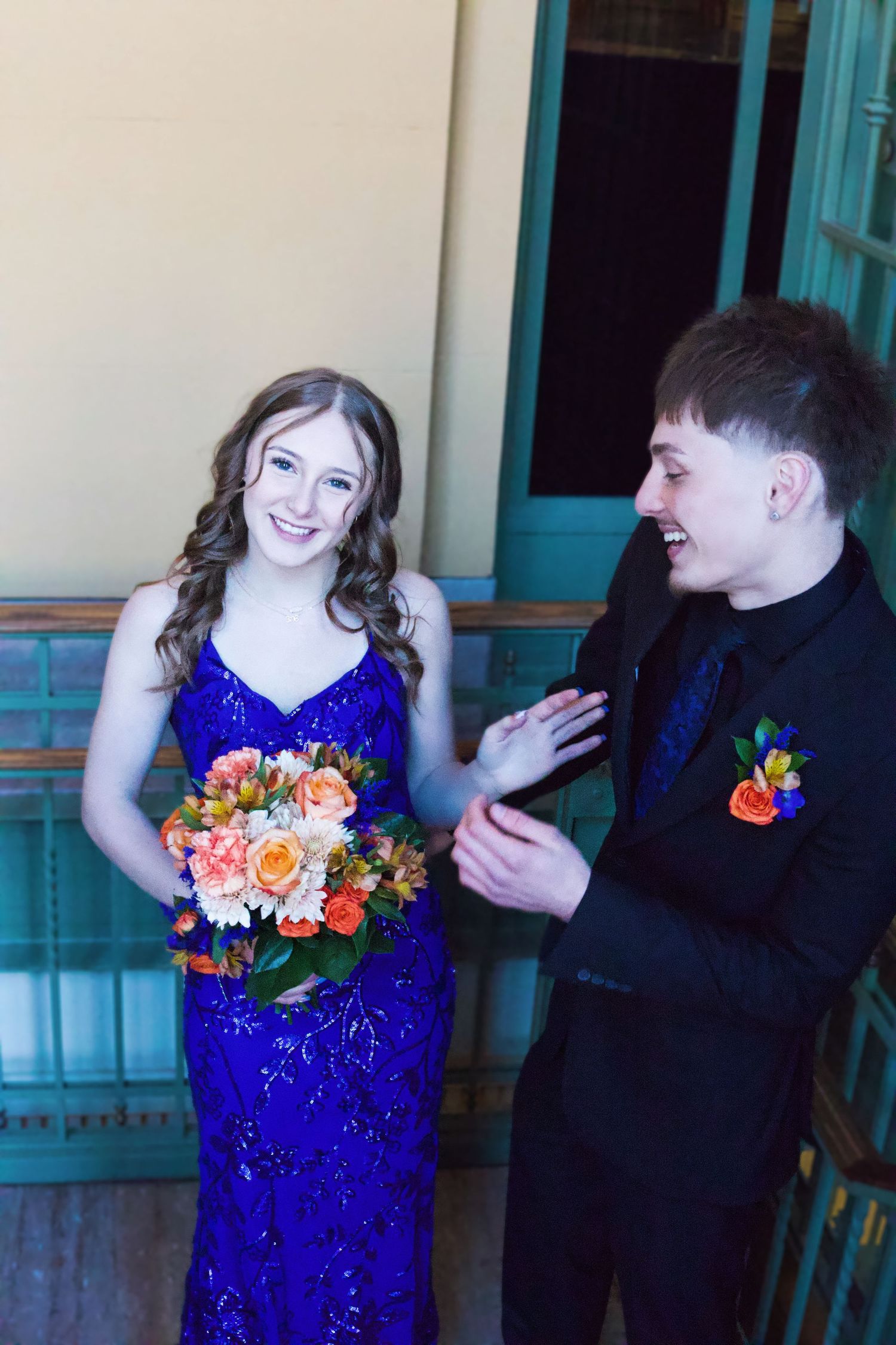 A smiling young couple in prom attire, girl in blue sequin gown with bouquet, boy in black suit with boutonniere.