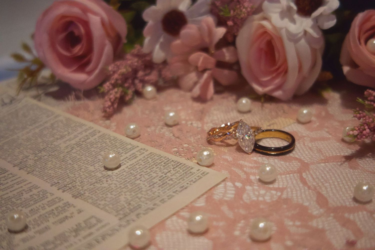 Gold diamond ring resting on pink lace with pearl beads, roses, and an open book in a romantic wedding flat lay.
