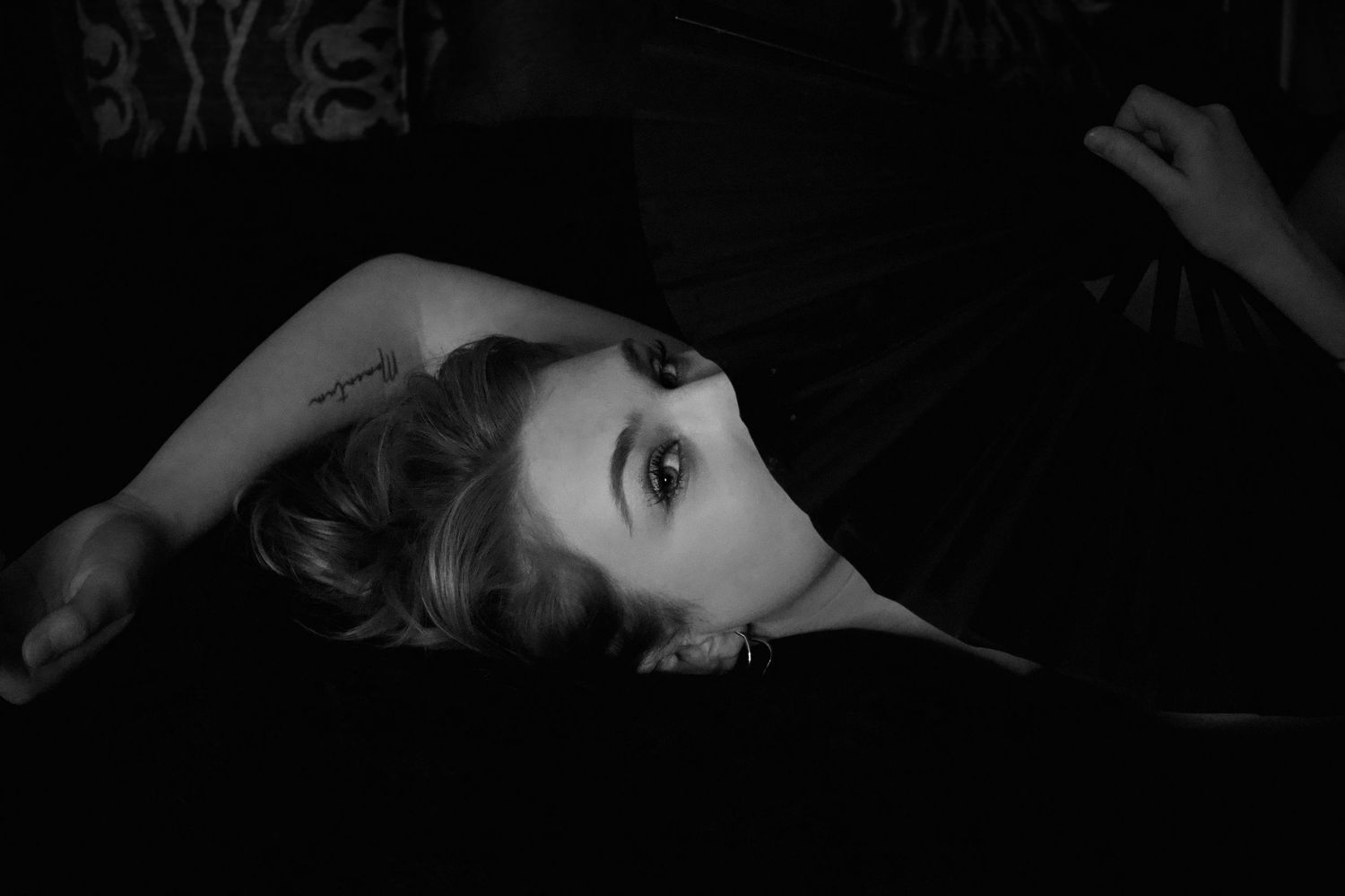 Black and white portrait of a woman lying down, gazing upward with dramatic lighting and dark background.