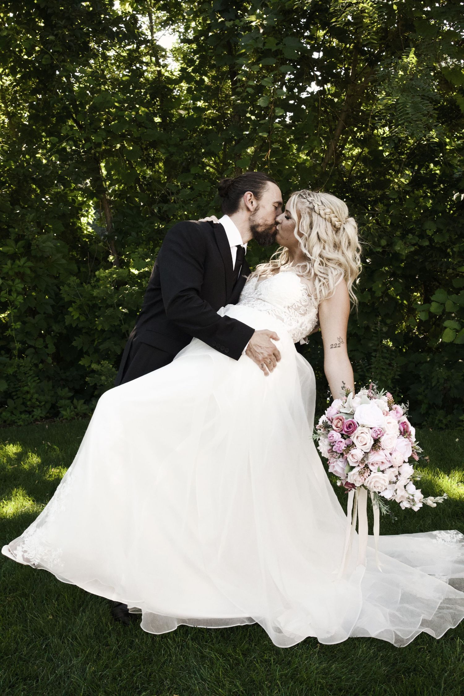 Bride and groom share a romantic kiss outdoors, surrounded by lush green trees, as bride holds pink floral bouquet.
