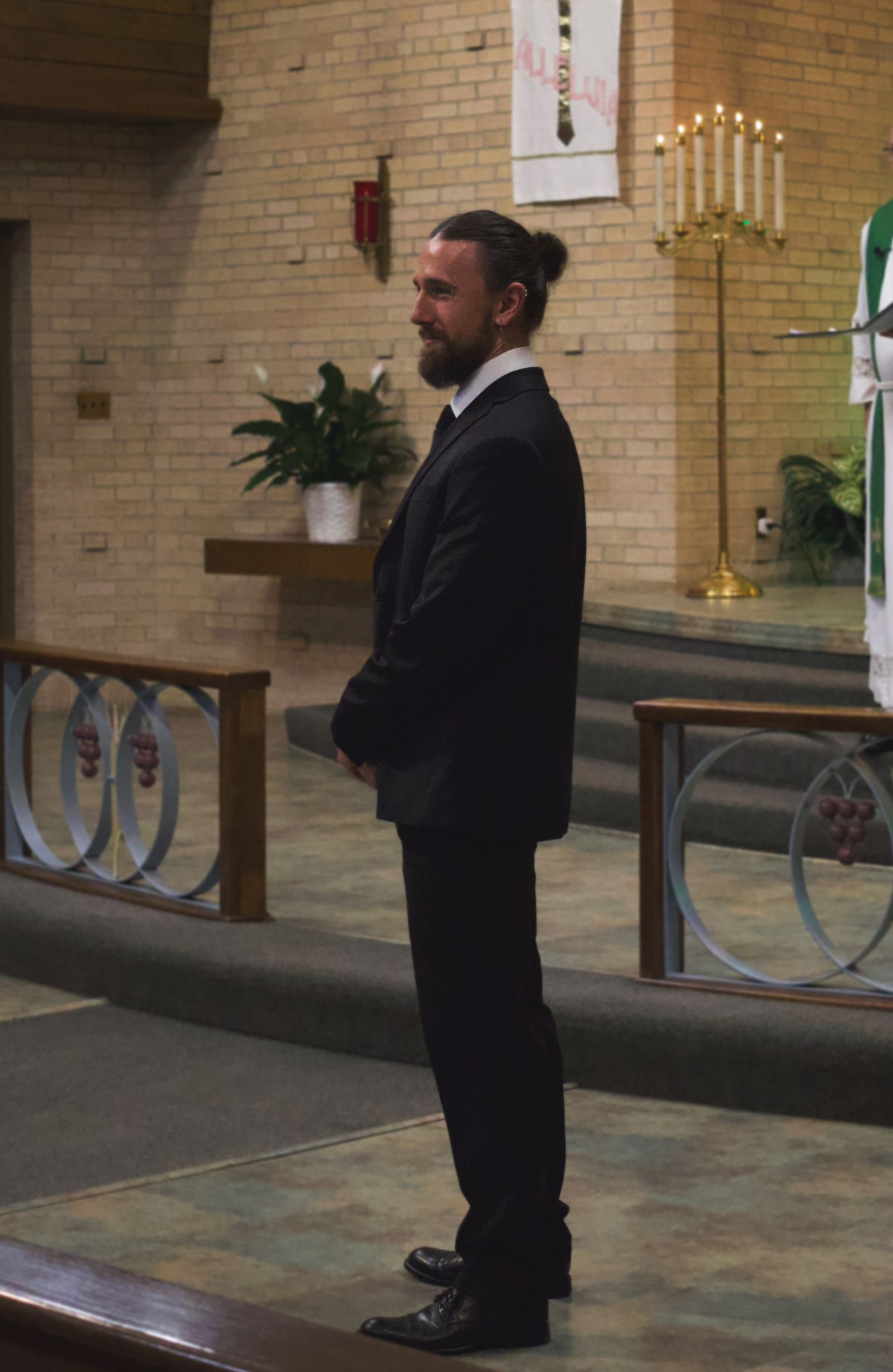 Man in black suit standing in church aisle with candles and altar in background.
