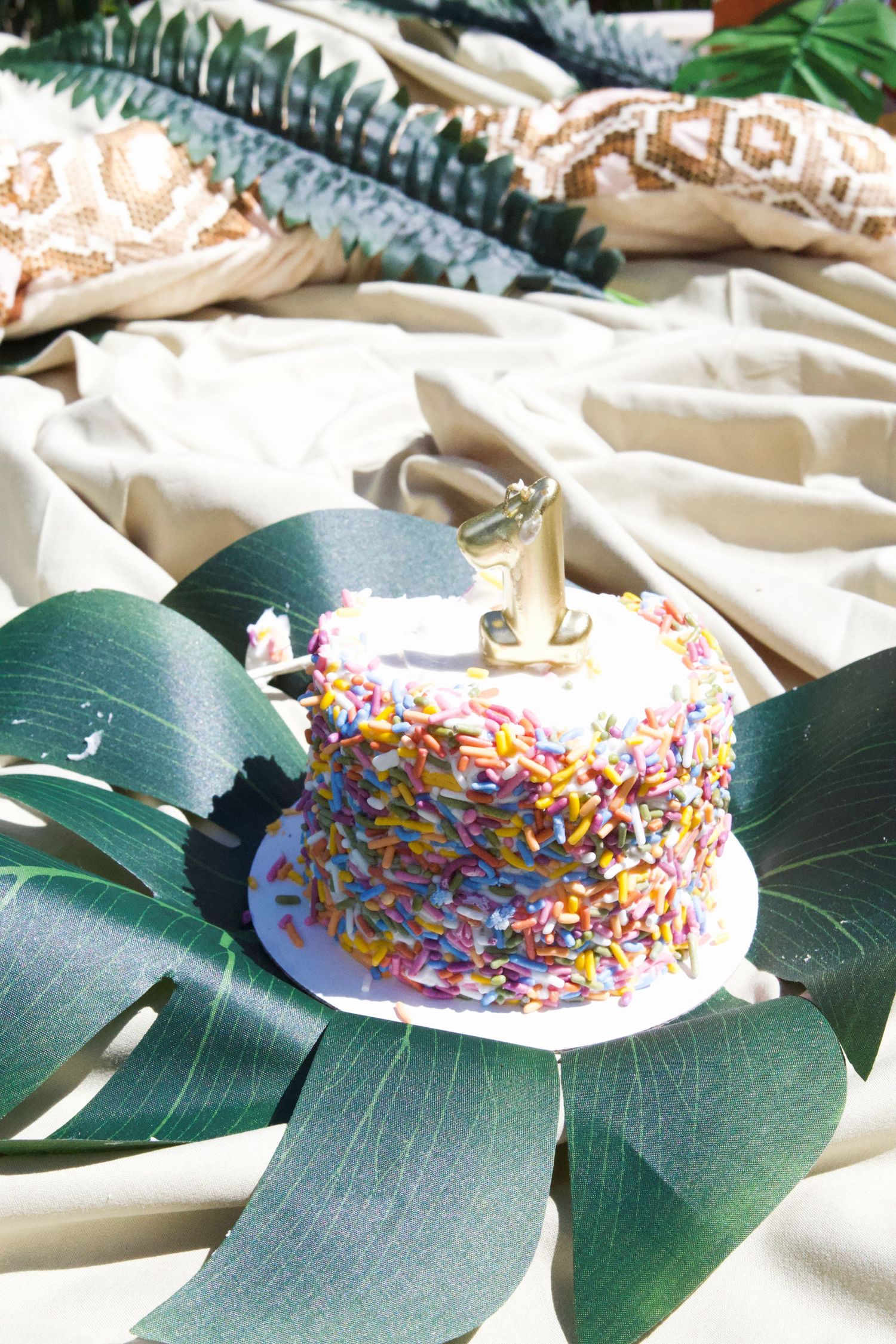 A colorful sprinkle-covered first birthday smash cake with gold number one topper on tropical green leaves.