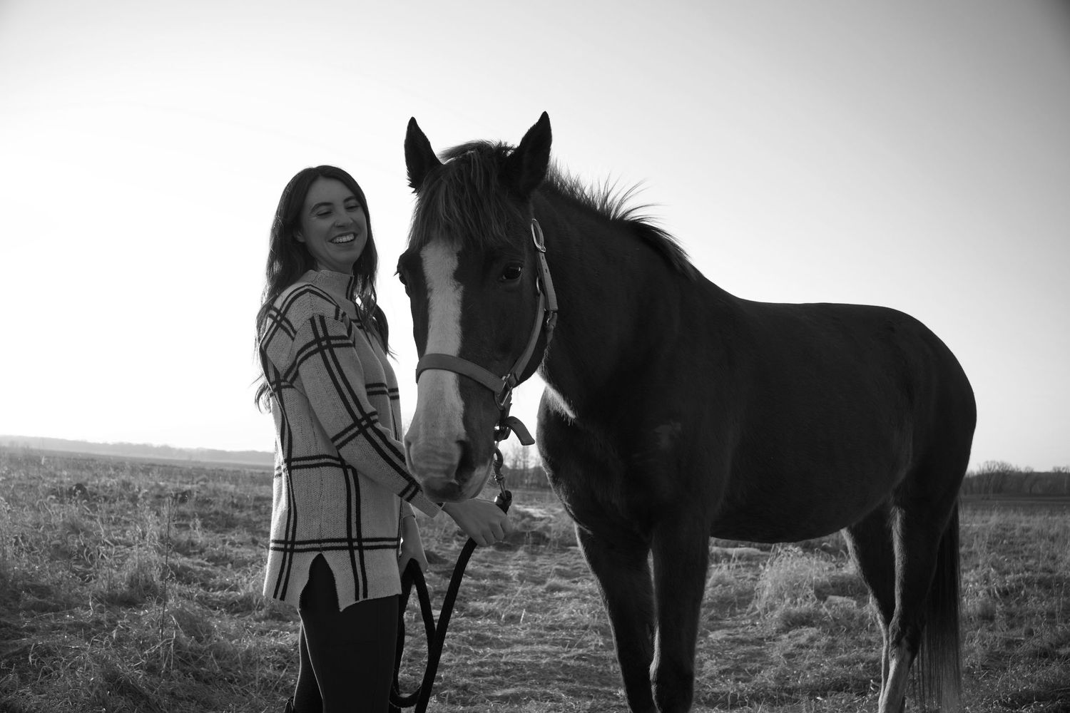 Black and white photo of a smiling woman standing beside a horse in an open field.