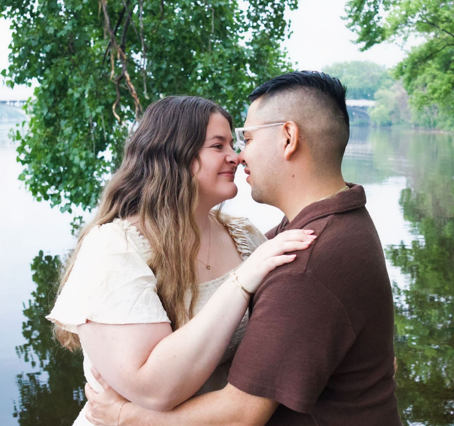 Couple sharing a romantic moment by a serene river, surrounded by lush green trees on a sunny day.