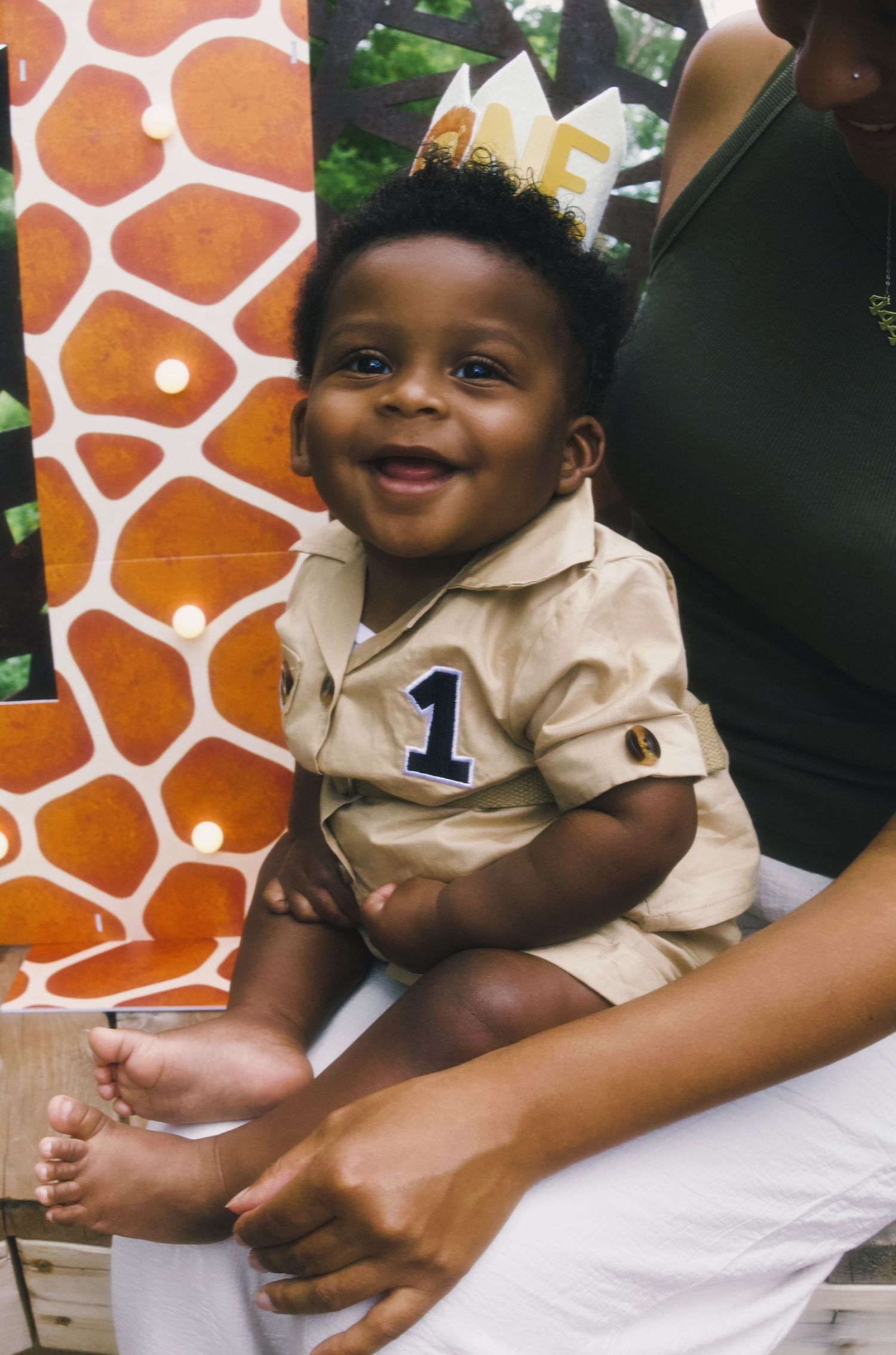 Smiling baby boy wearing a number 1 outfit and birthday crown sits happily in front of a giraffe print backdrop.