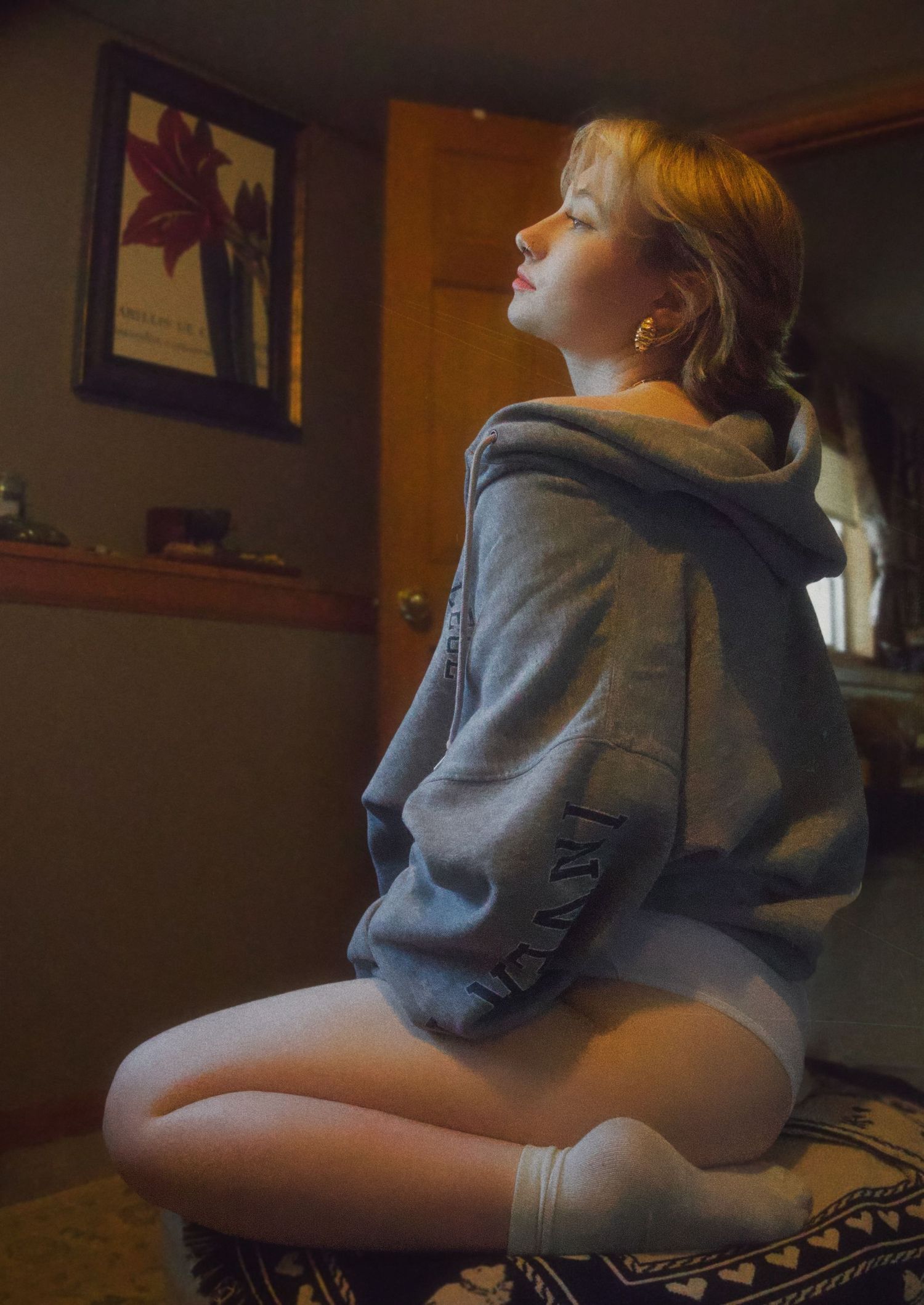 Young woman in gray hoodie and white shorts sitting cross-legged on ottoman in warmly lit room.