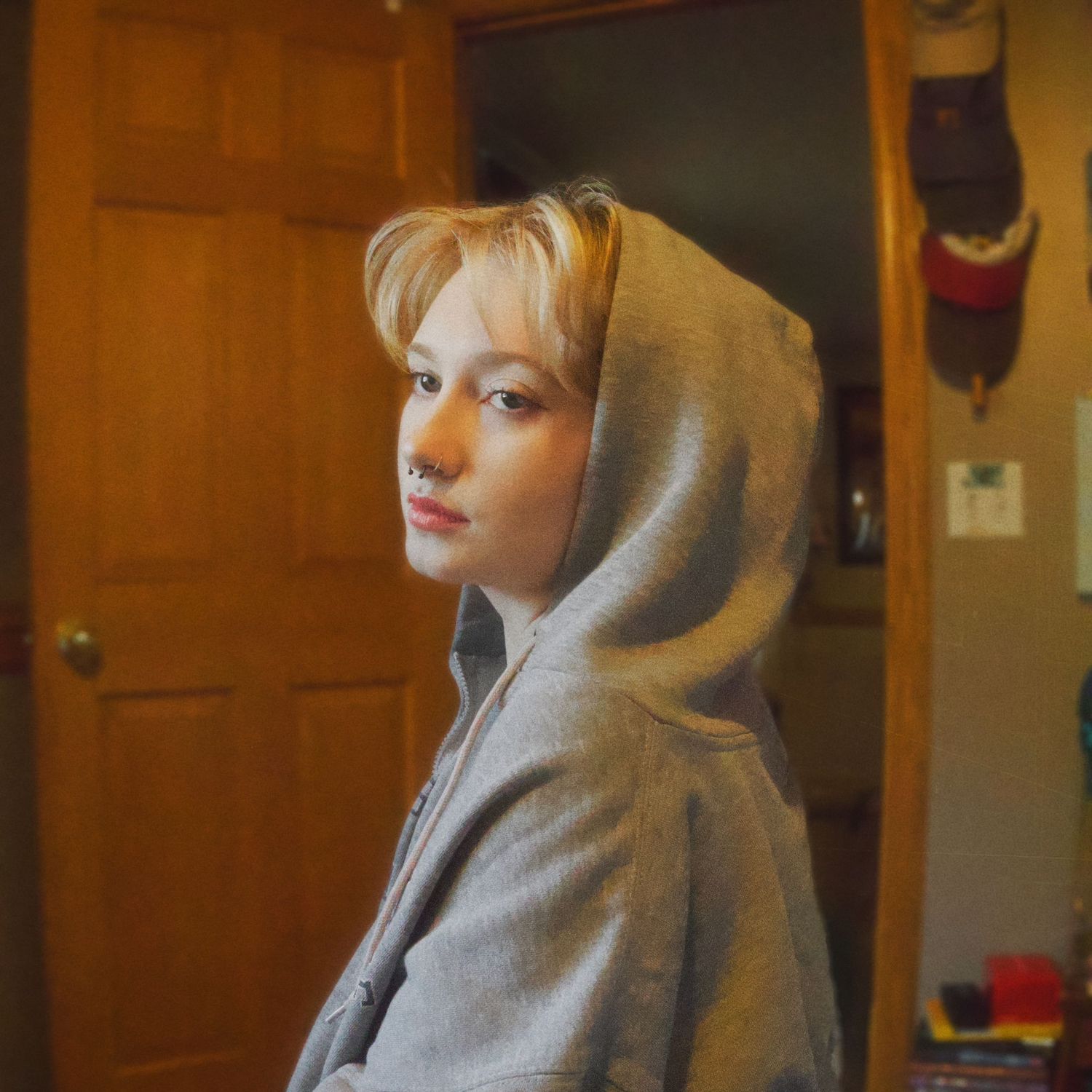 Young woman with blonde hair wearing a gray hoodie, standing indoors near a wooden door with a nose piercing.