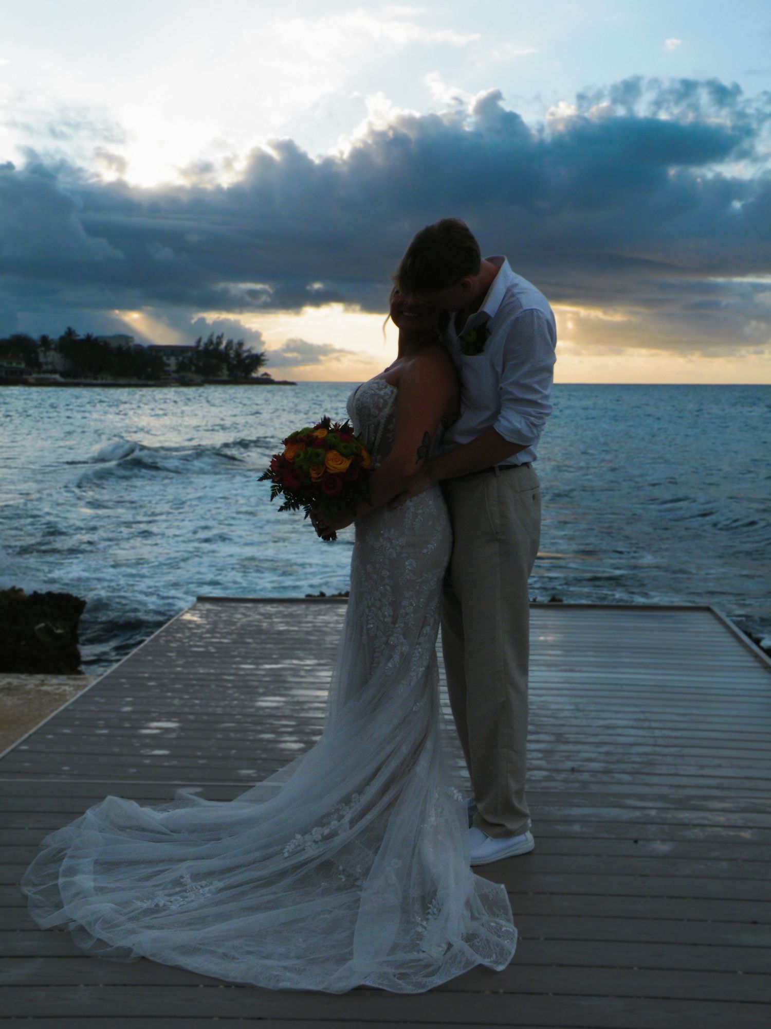 Newlywed couple sharing a romantic kiss at sunset on a wooden dock by the ocean, bride in lace gown holding bouquet.