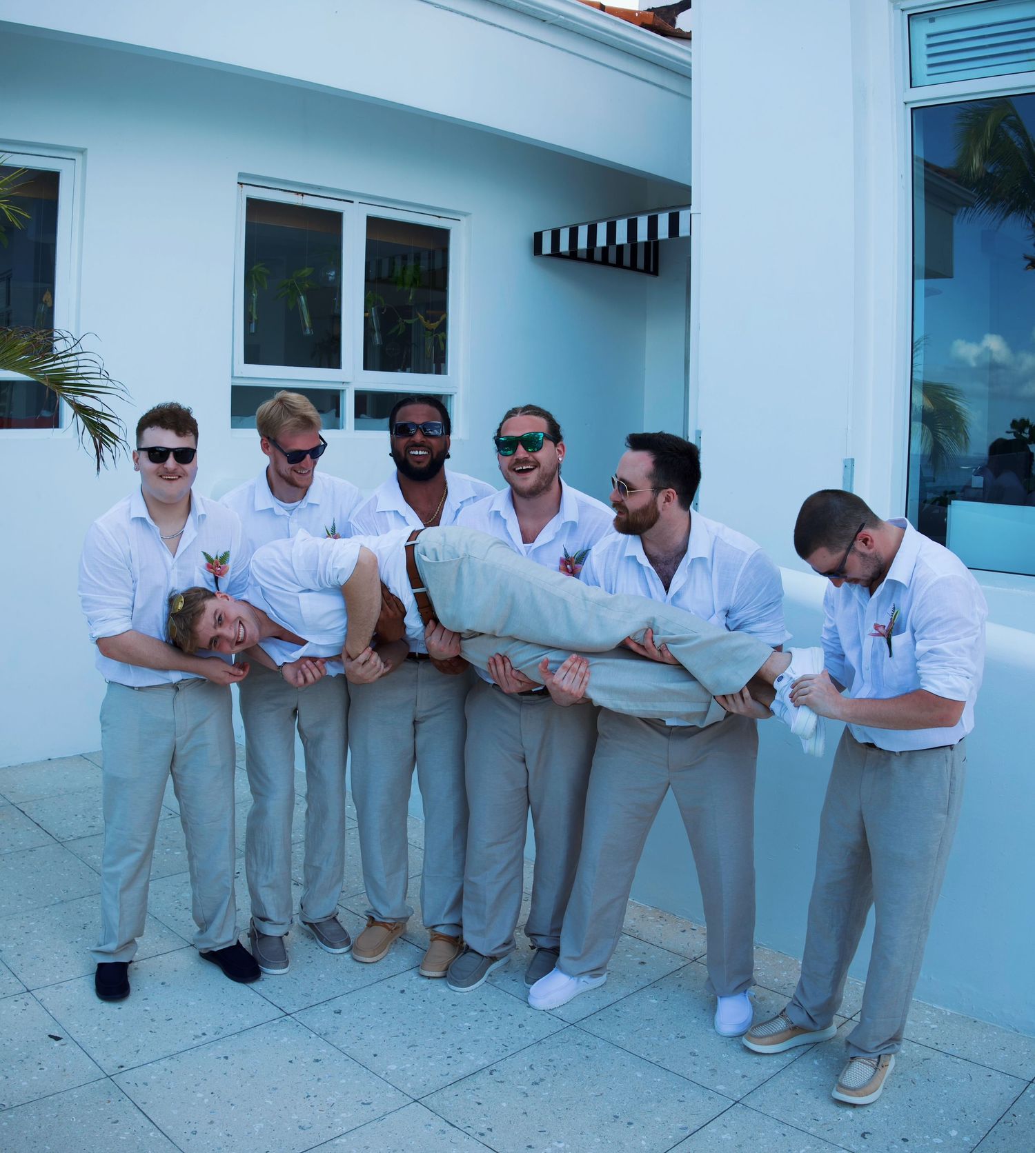Group of groomsmen in white shirts and khaki pants playfully carrying groom at outdoor wedding venue.