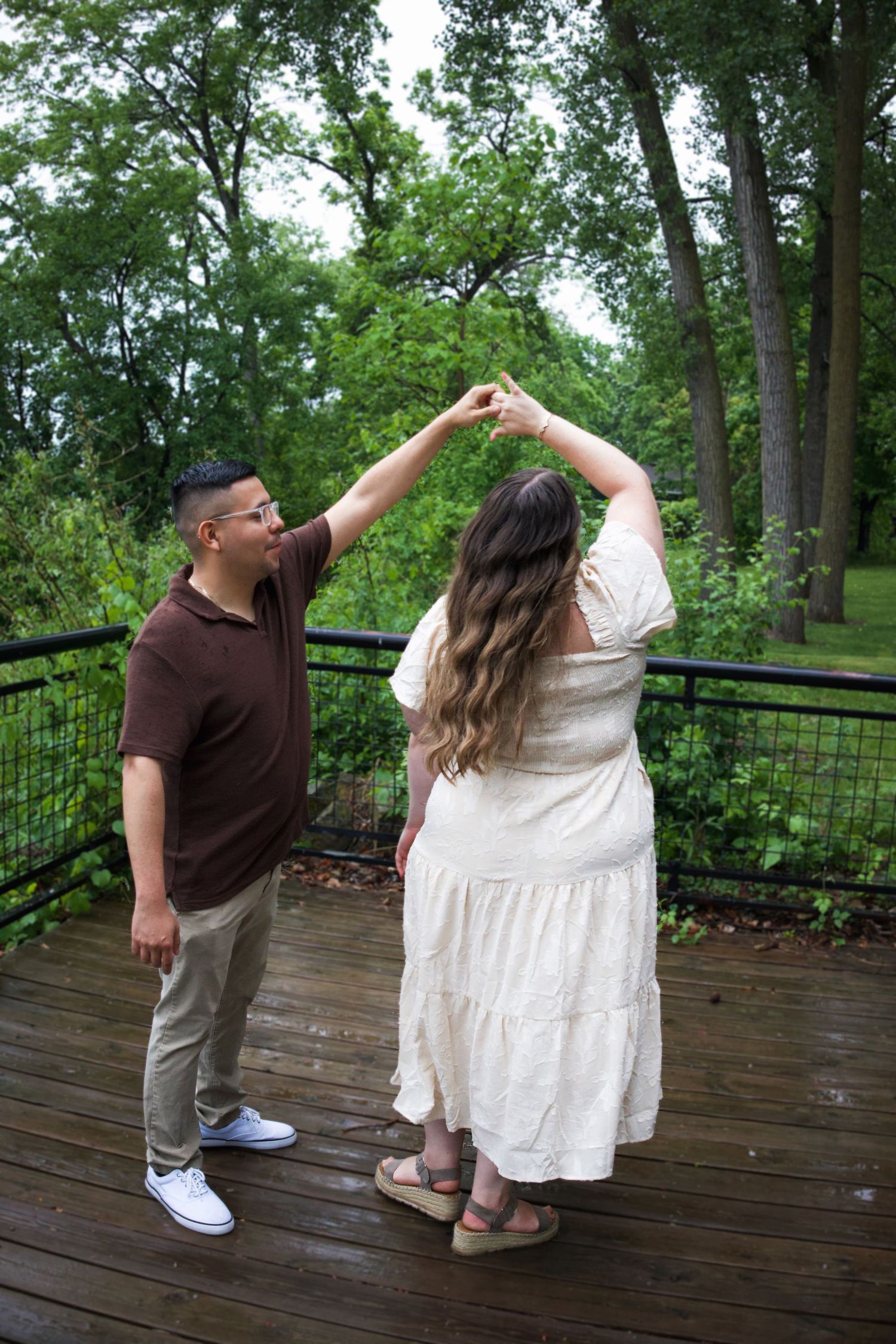 Couple dancing on a wooden deck surrounded by lush green trees, man in brown shirt twirling woman in white dress.