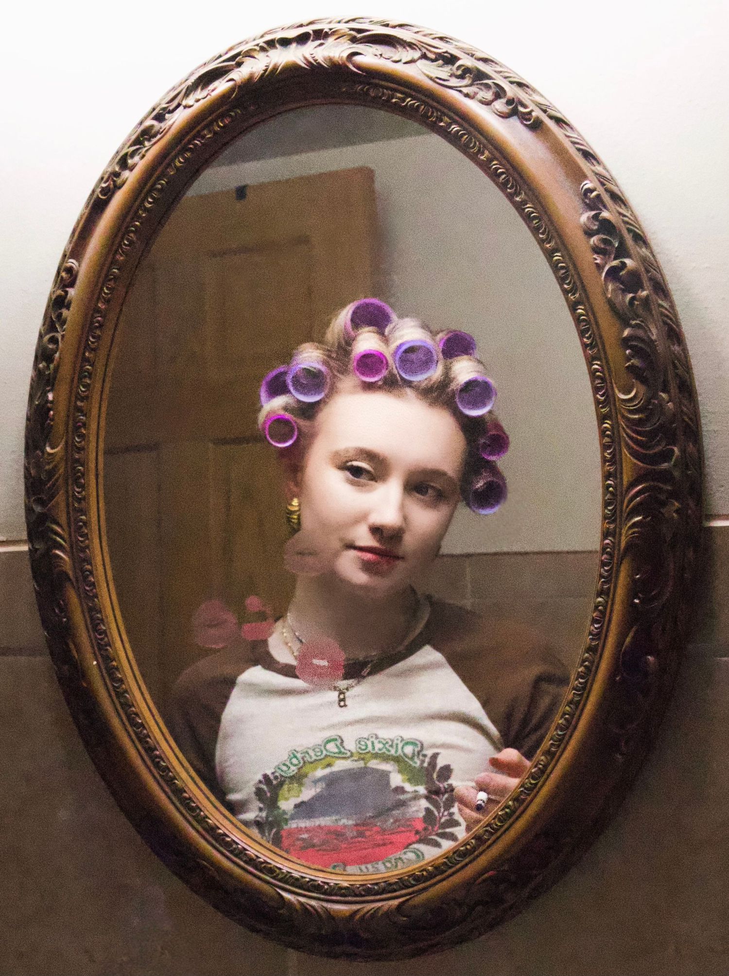 Young woman with purple hair rollers gazes into an ornate oval gold-framed mirror in a vintage-style setting.