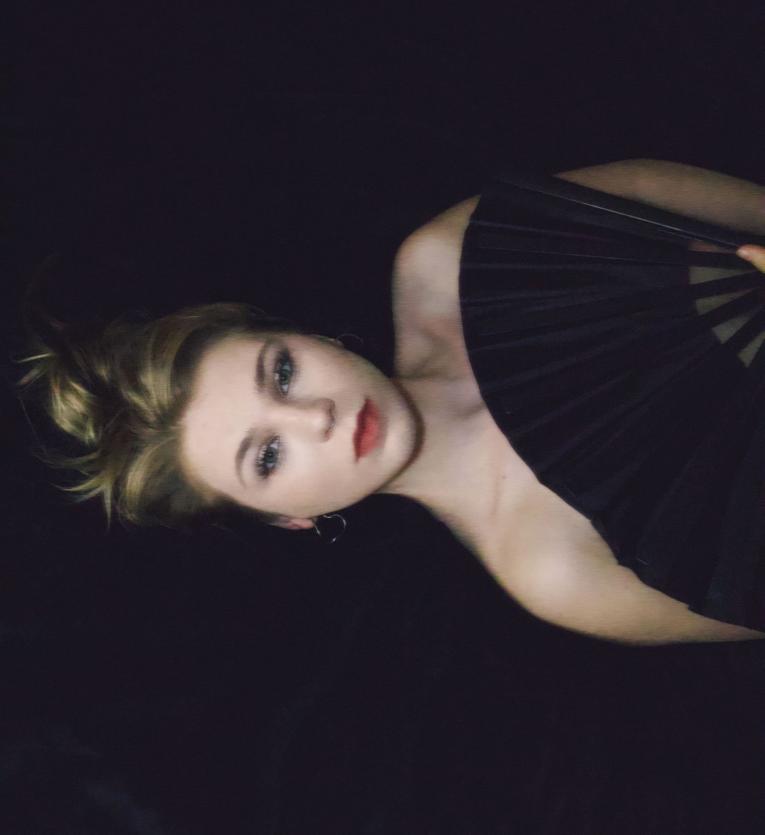 Young woman in black dress posing dramatically against dark background with elegant updo hairstyle.