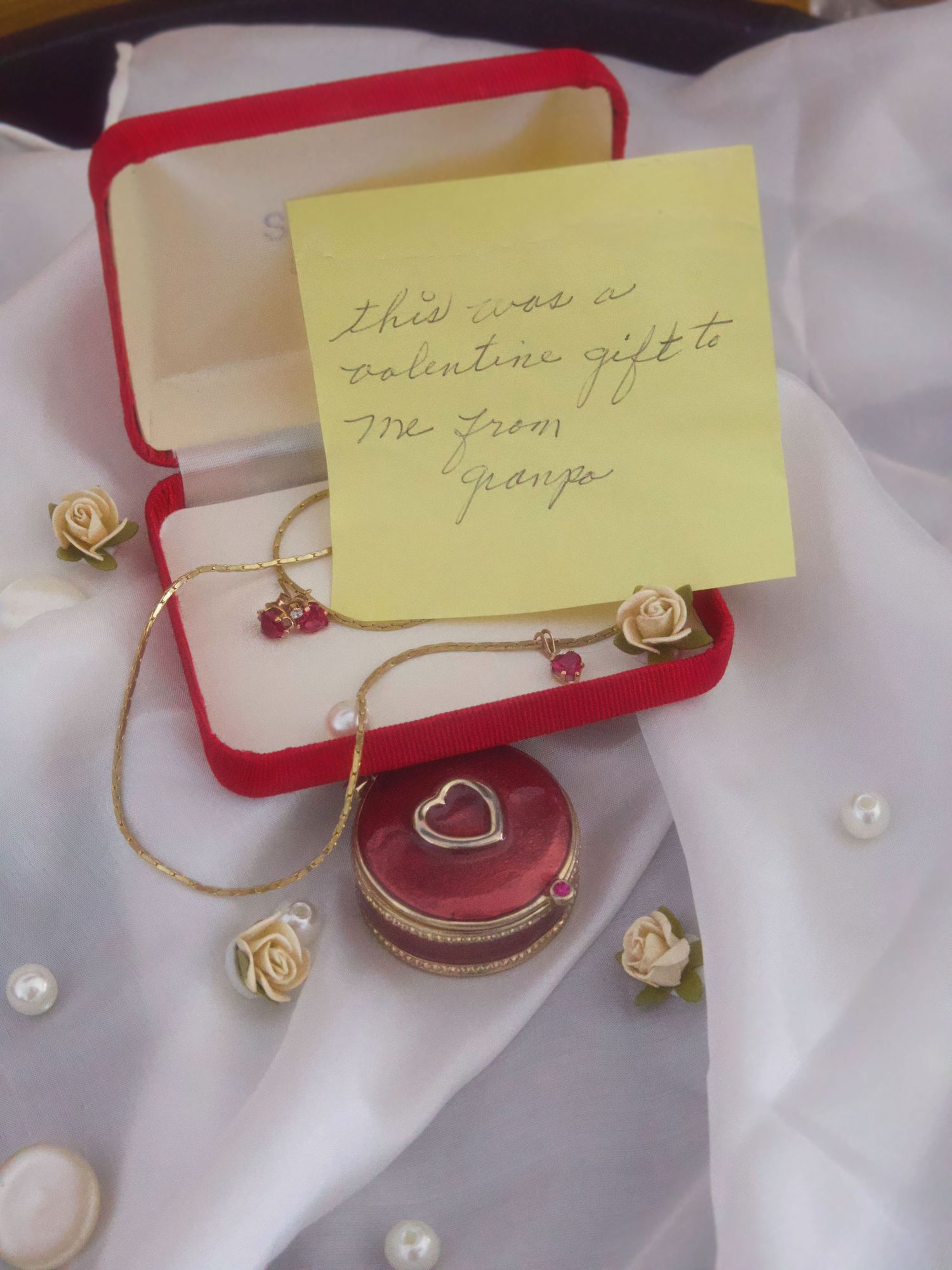 Red jewelry box with gold heart necklace and earrings, a sticky note reading valentines gift from grandpa, on white fabric.