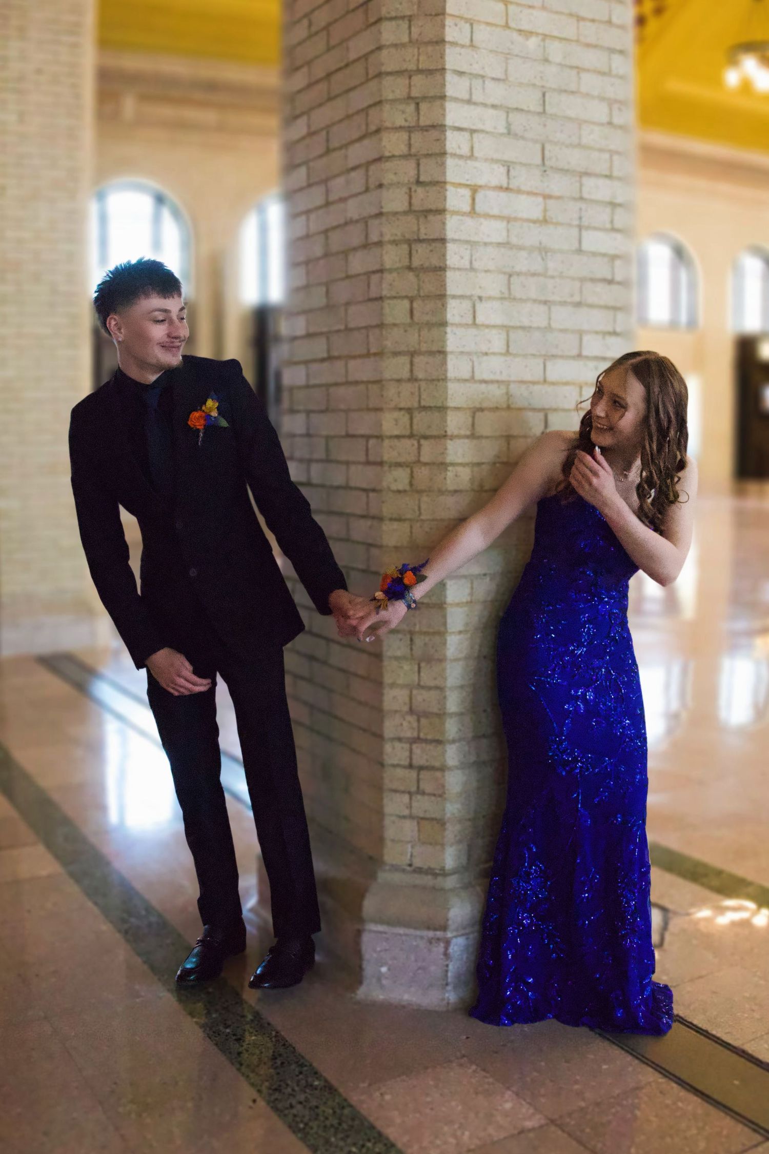 A young couple in prom attire, man in black suit and woman in royal blue sequin gown, holding hands by a brick column.