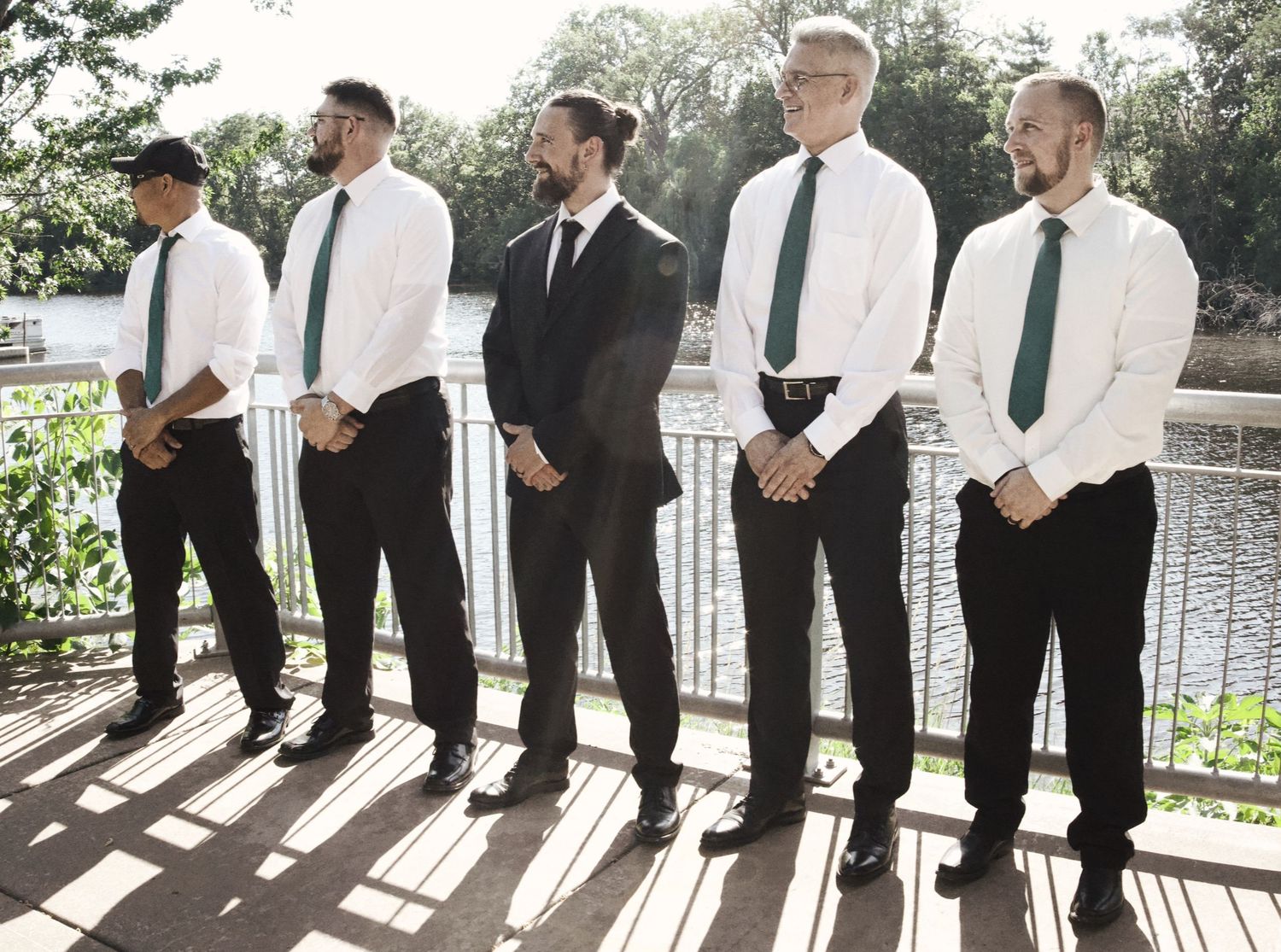 Five groomsmen in white shirts and green ties stand on a sunlit outdoor deck near a scenic waterway.