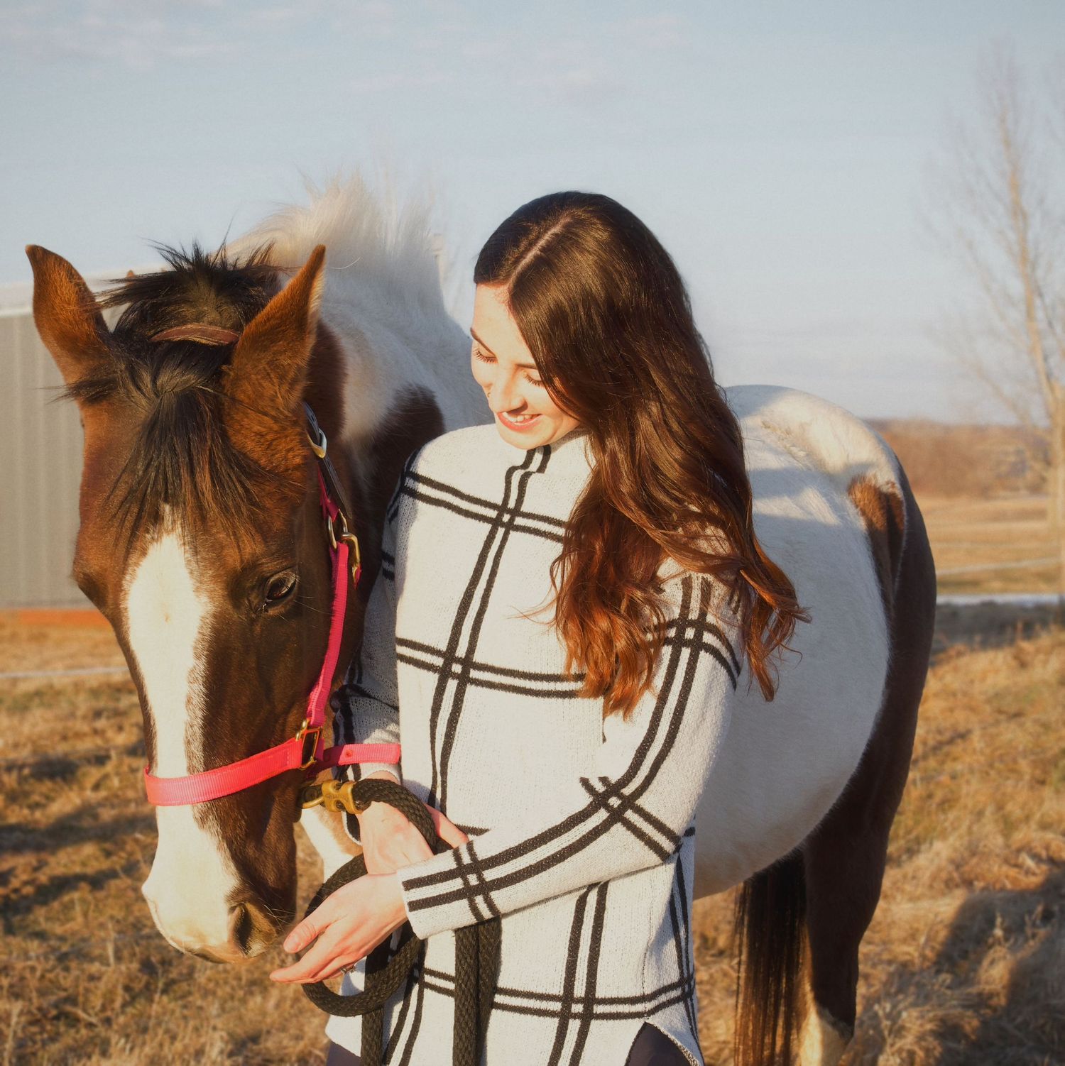 Young woman with long brown hair smiling beside a brown and white paint horse wearing a red halter outdoors.