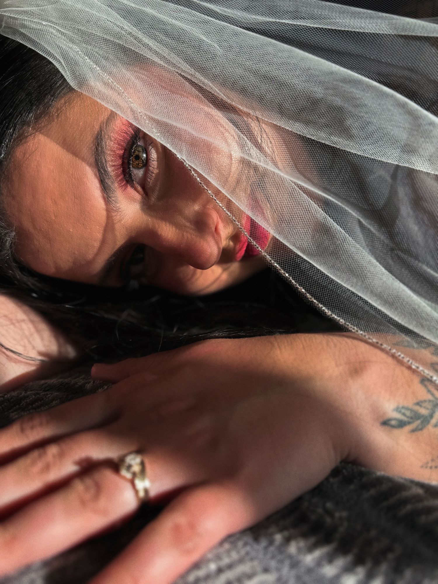 Woman with dramatic pink eye makeup and tattoos poses under sheer white veil with silver rings on hand.