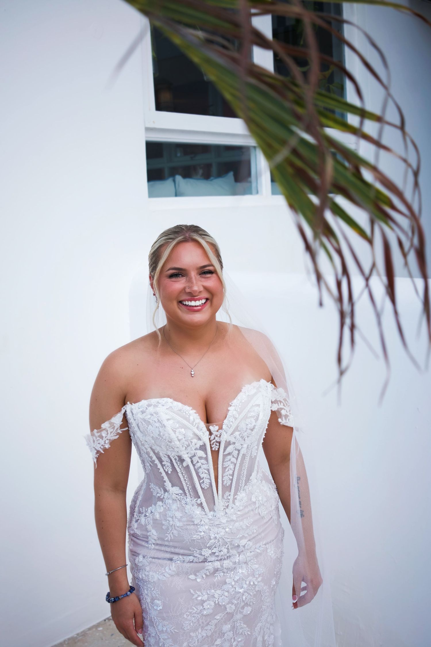 Smiling bride in off-shoulder lace wedding gown with deep V-neckline poses outdoors near white wall and tropical palm.