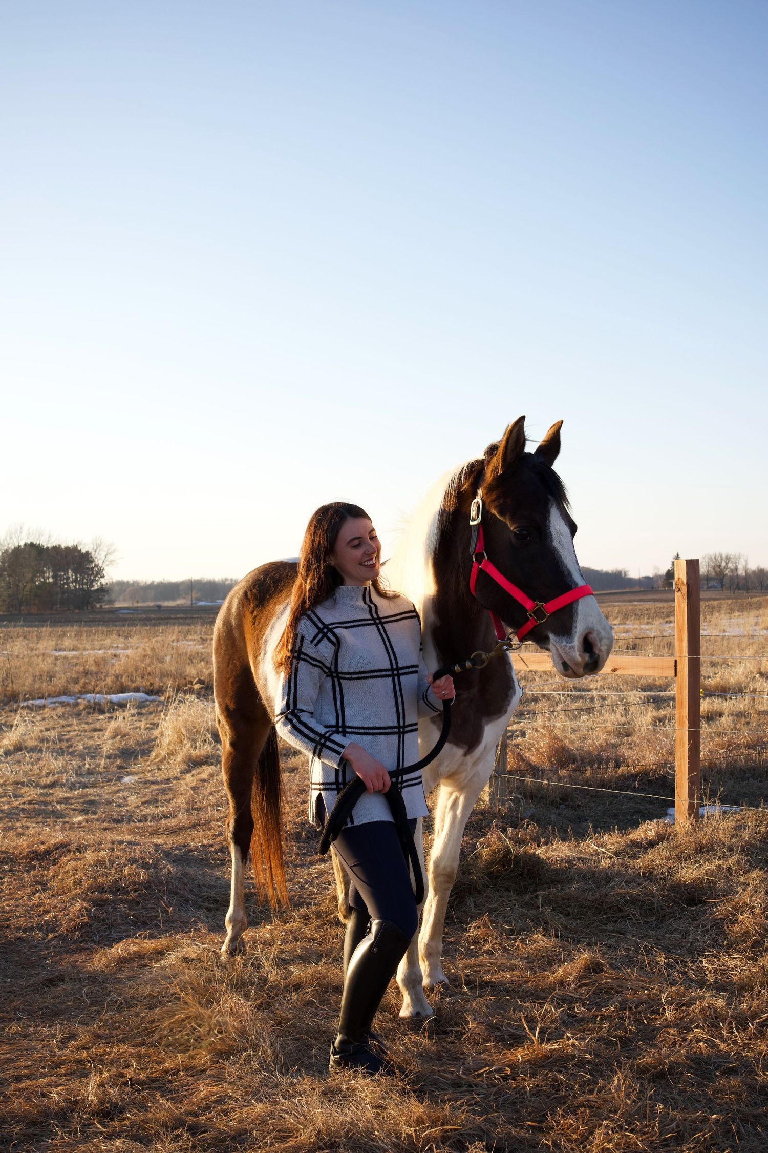 Young woman in plaid sweater smiling beside a brown and white horse with red halter in a sunny rural field.