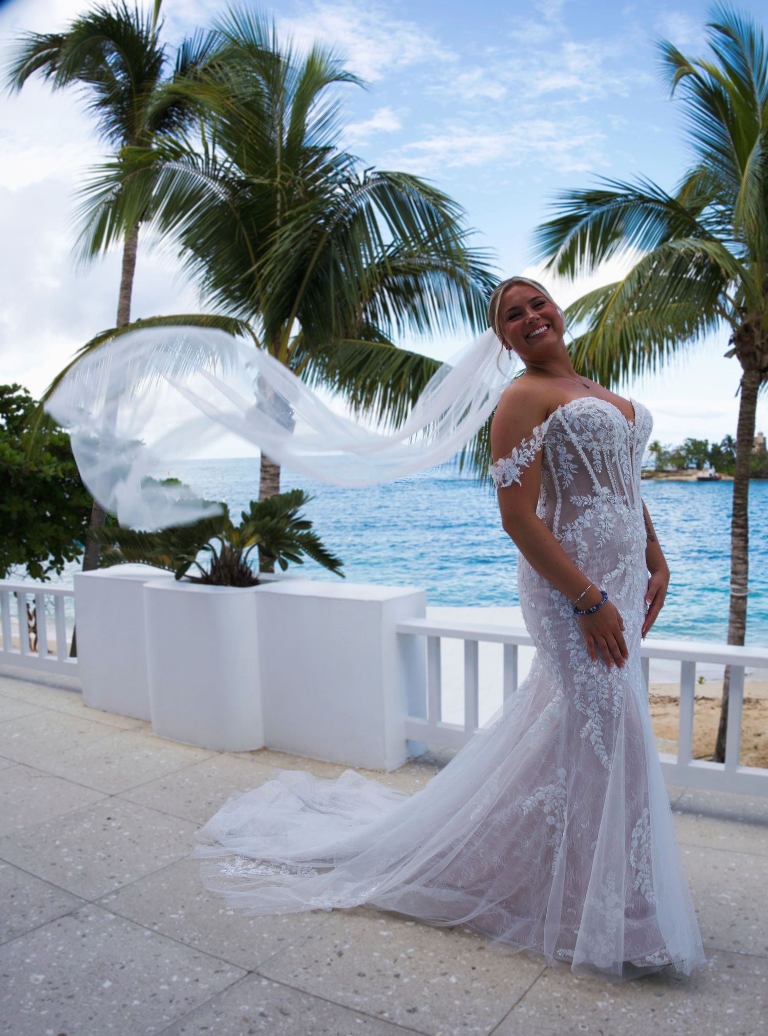 Bride in lace mermaid wedding gown with flowing veil on tropical beach terrace with palm trees and ocean backdrop.