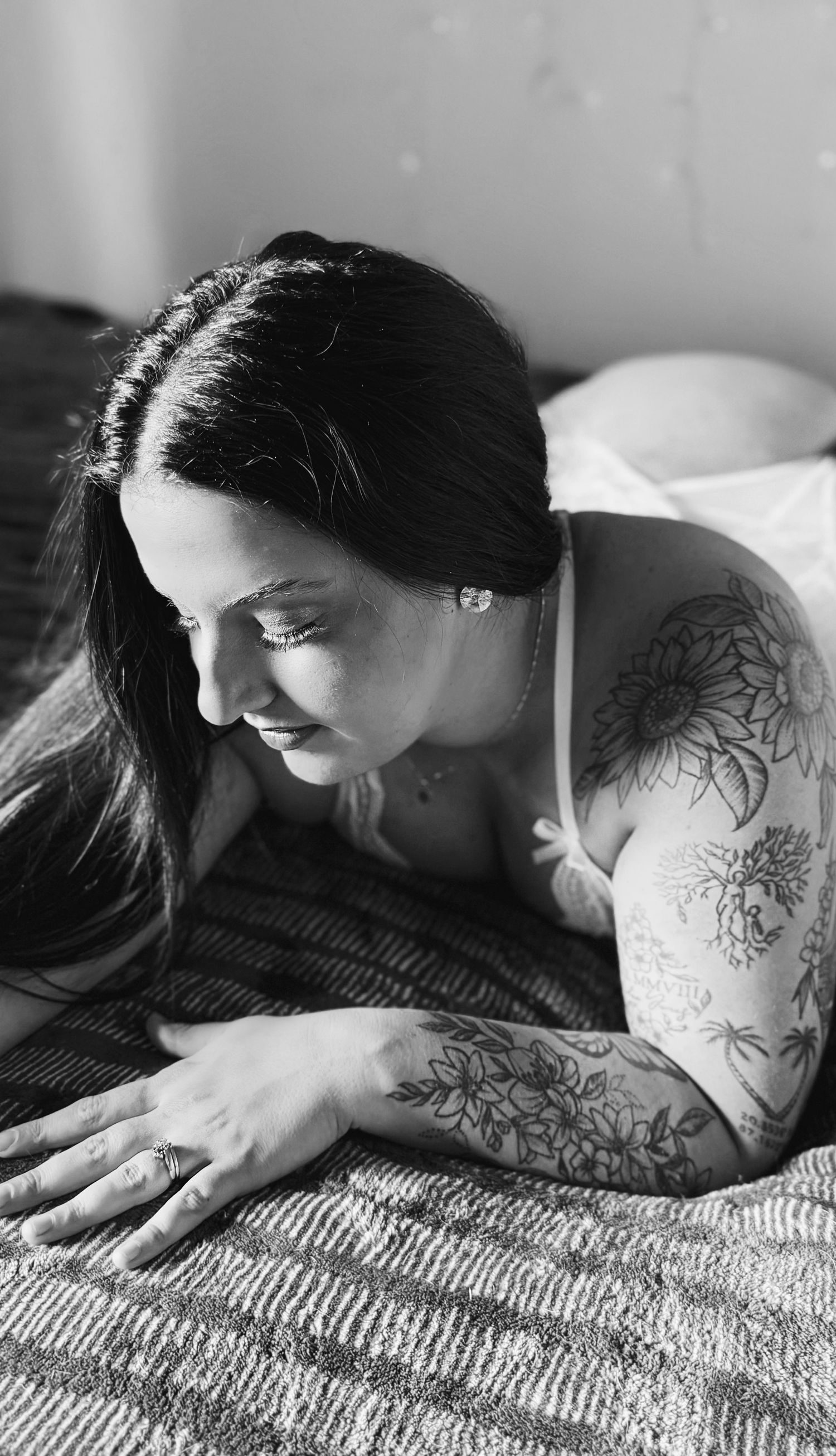 Black and white photo of a tattooed woman lying on a bed, looking down thoughtfully.