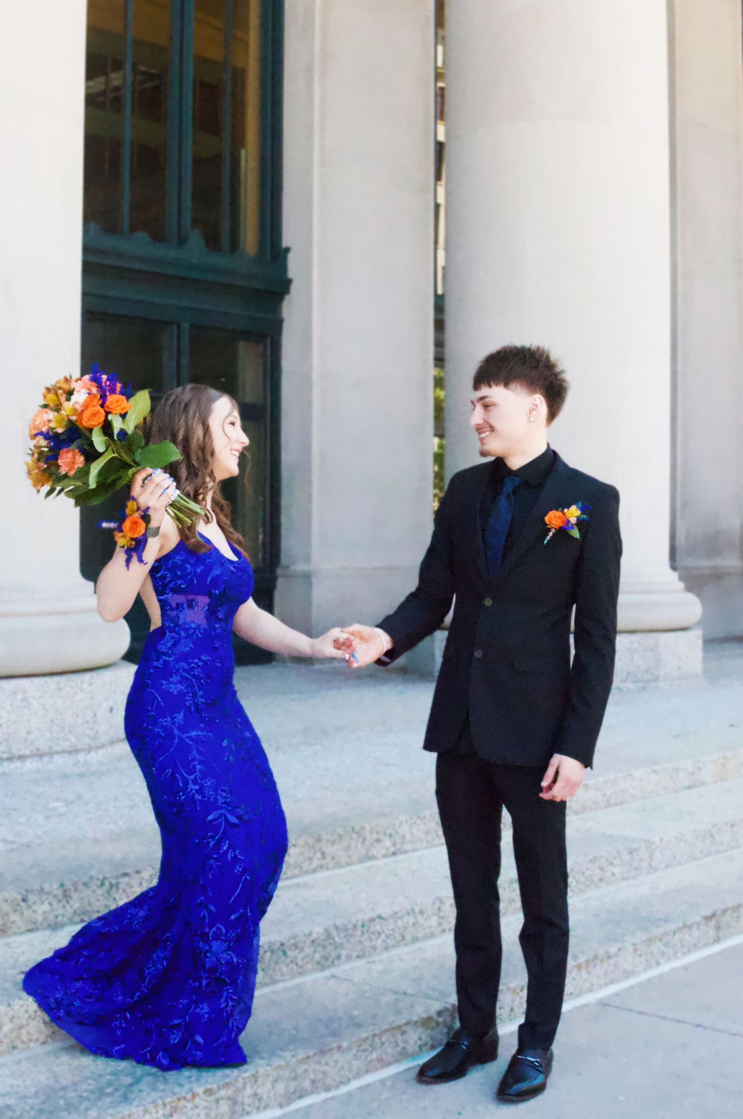 A couple in formal attire, blue gown and black suit, hold hands on steps with colorful bouquet.