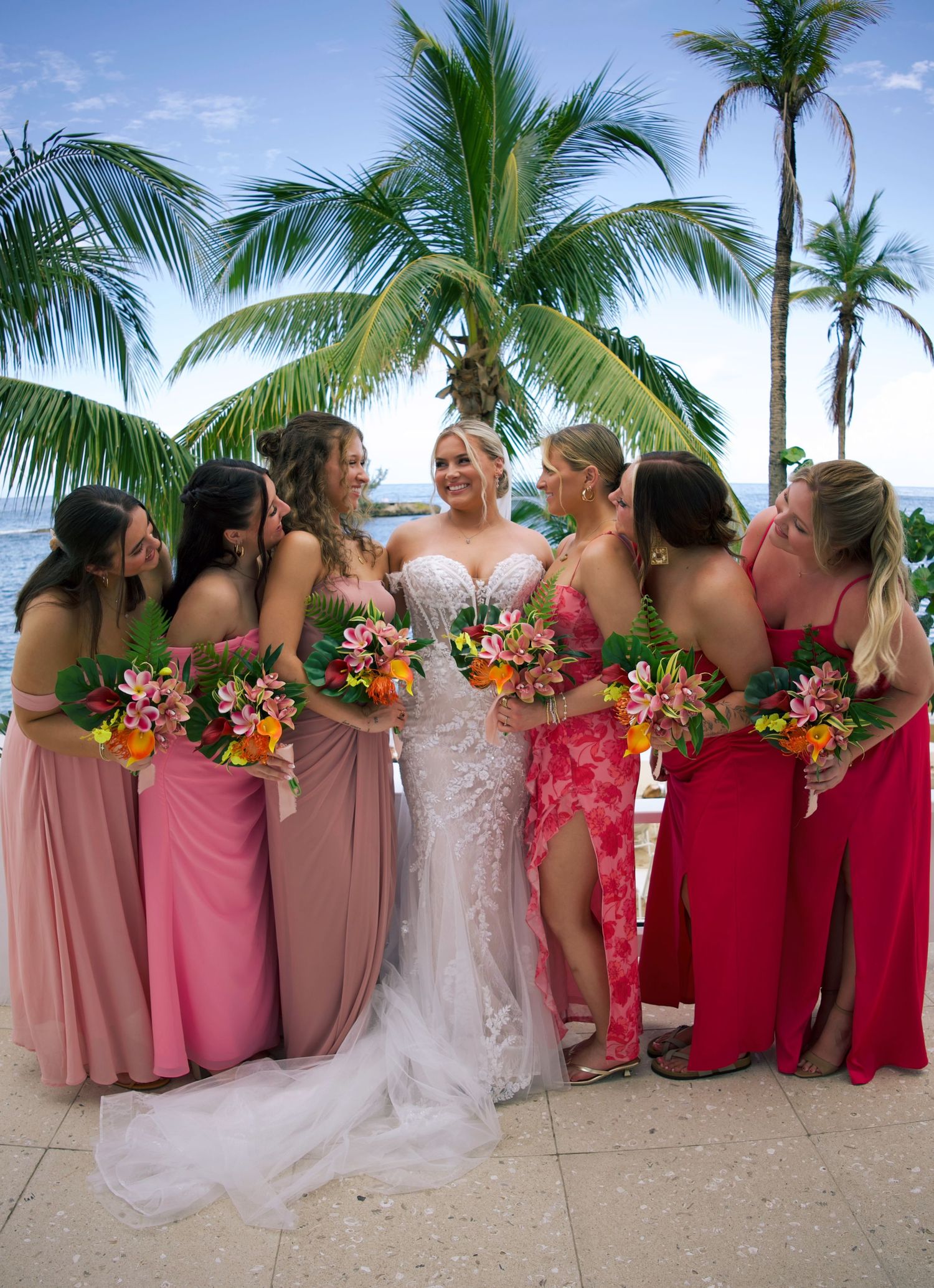 Bride in white lace gown surrounded by bridesmaids in pink dresses holding tropical bouquets near palm trees at beach wedding.
