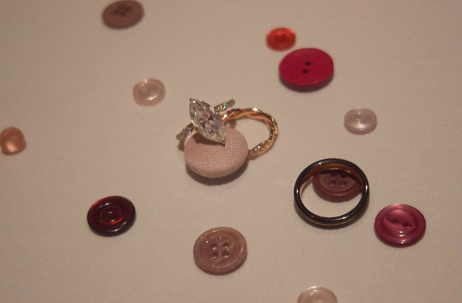 Gold diamond ring and black band ring surrounded by scattered pink and red buttons on a white surface.