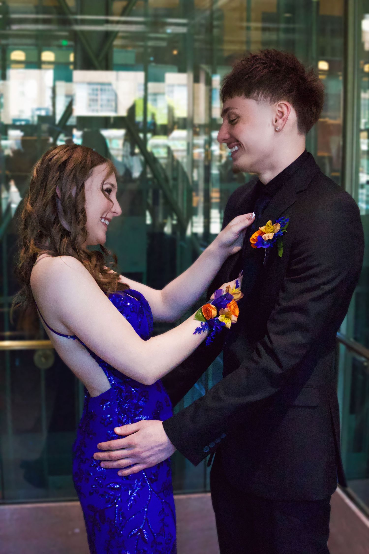 Young couple in prom attire, girl in blue sequin dress and boy in black suit with boutonniere, smiling at each other.