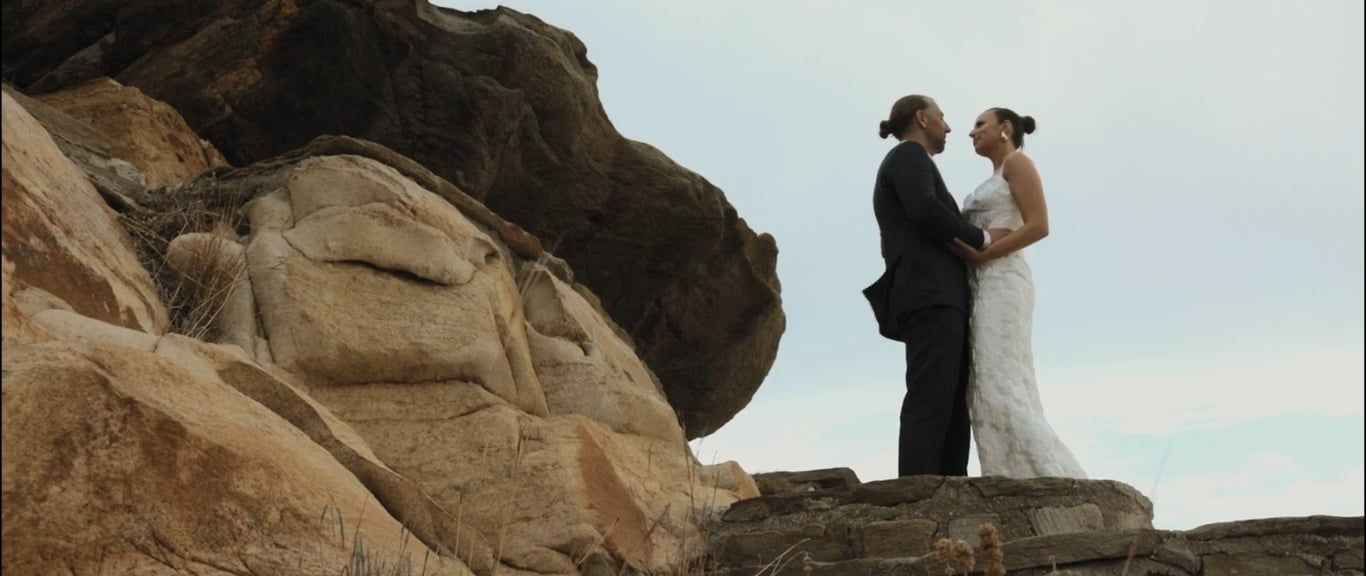 A couple in formal wedding attire stands atop dramatic red rock formations in an outdoor desert landscape.