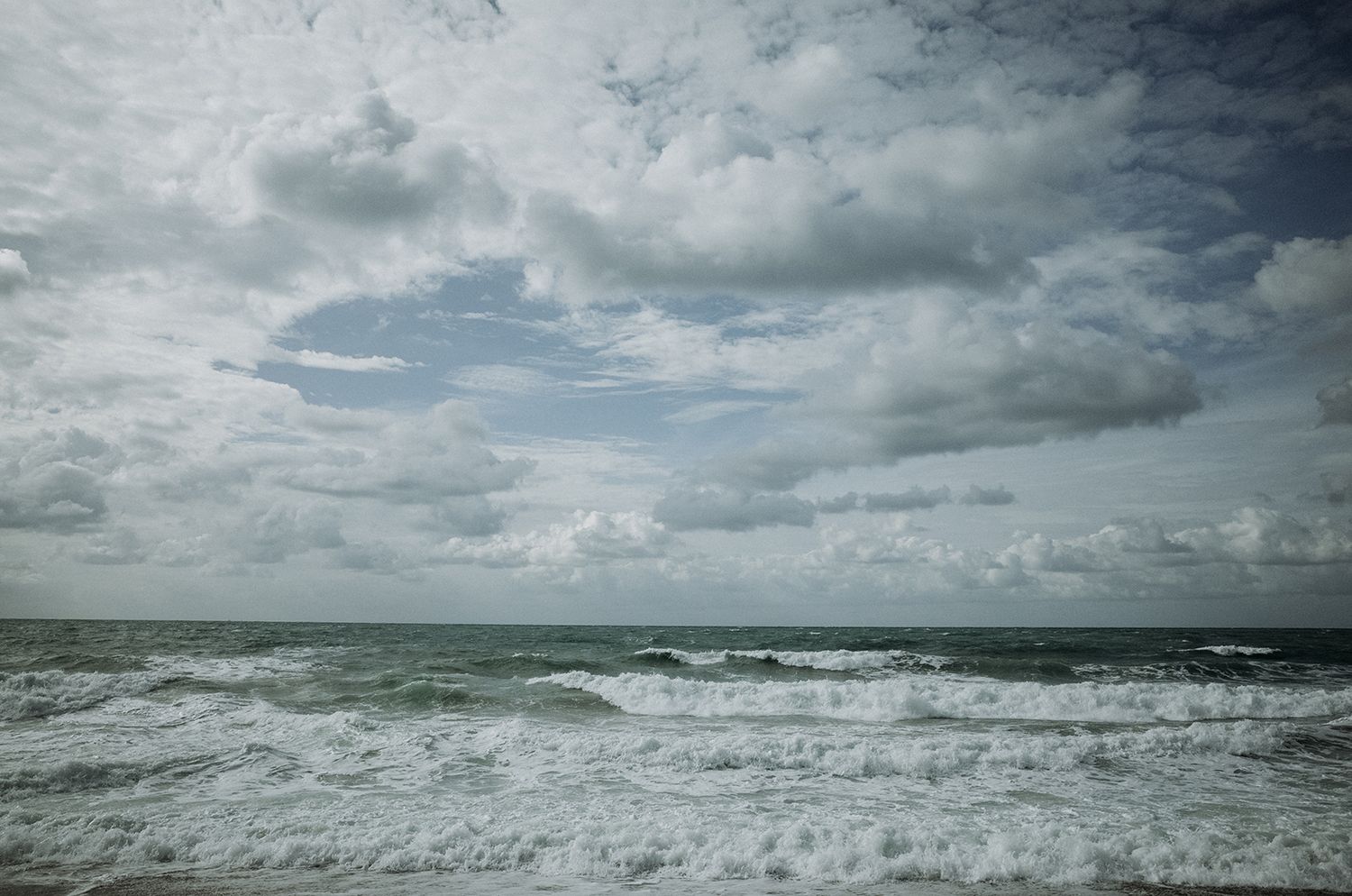 Dramatic seascape panorama showing stormy ocean waves under moody cloudy skies along a rugged coastline.