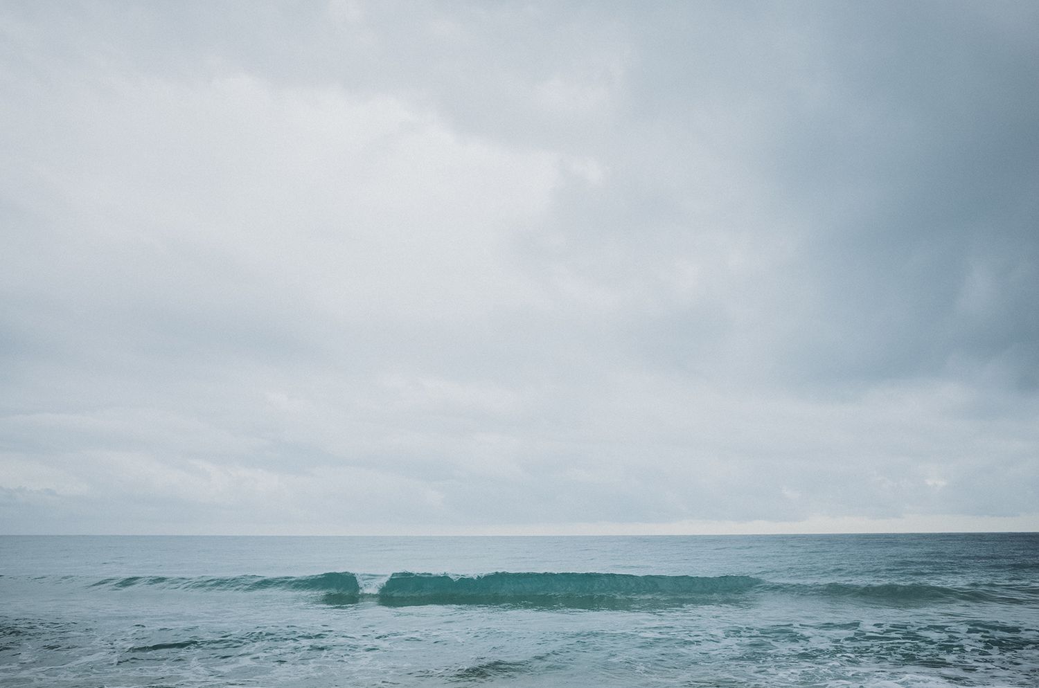 A turquoise ocean wave crests under an overcast sky with stormy clouds looming above the horizon.