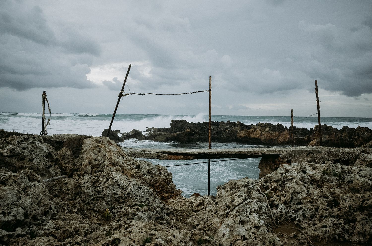 Wooden poles stand among rocky tide pools along a moody coastline under stormy gray clouds.