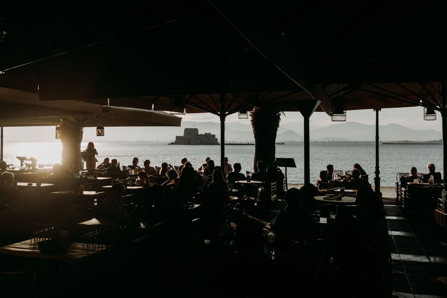 Silhouetted waterfront restaurant scene at sunset with ocean view.