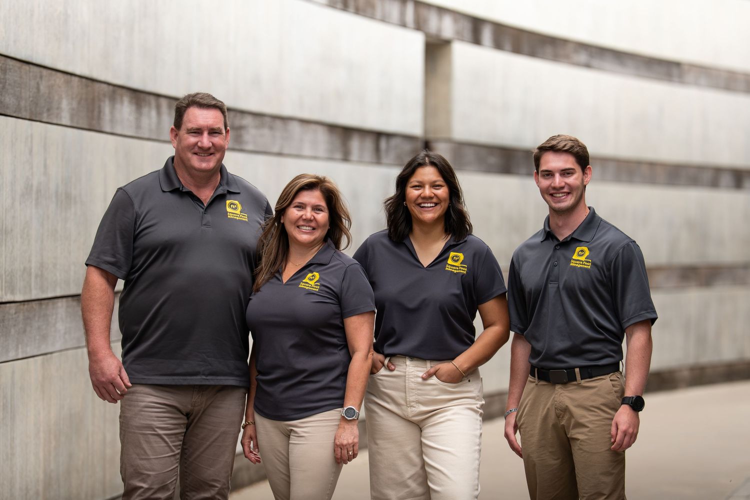 Four colleagues in matching dark polo shirts with gold logos smile together outdoors against a concrete wall.