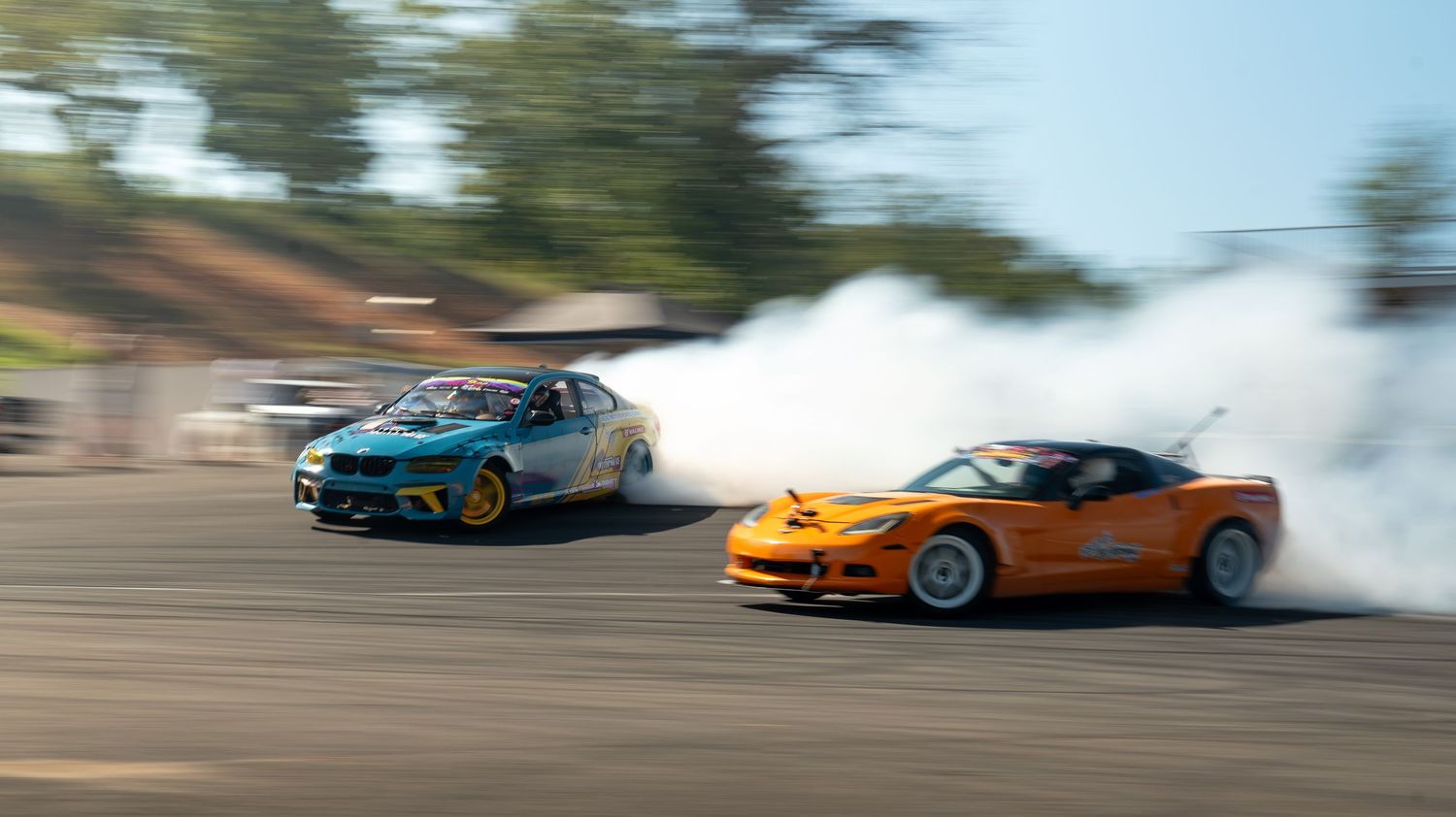 Modified sports cars drifting in formation on a race track with smoke trailing behind them.