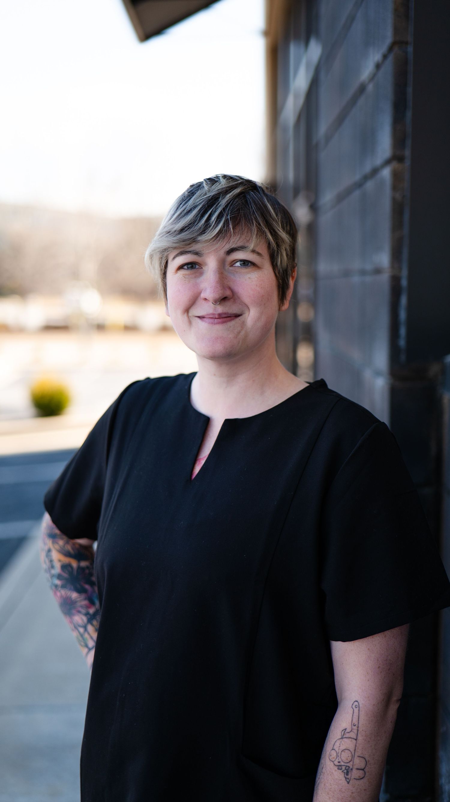 Person in black shirt with tattoos standing near a window.