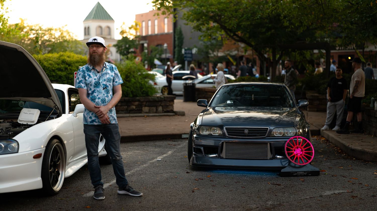 Panoramic shot of car meet location with modified vehicles and trees in evening light.