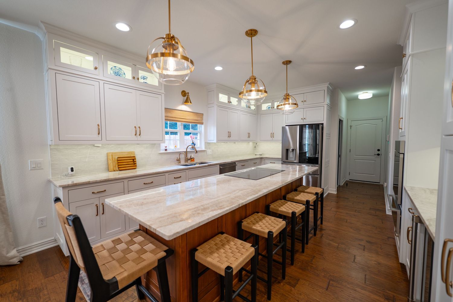 White kitchen with gold pendant lights, marble island, bar stools, and stainless steel appliances on dark hardwood floors.