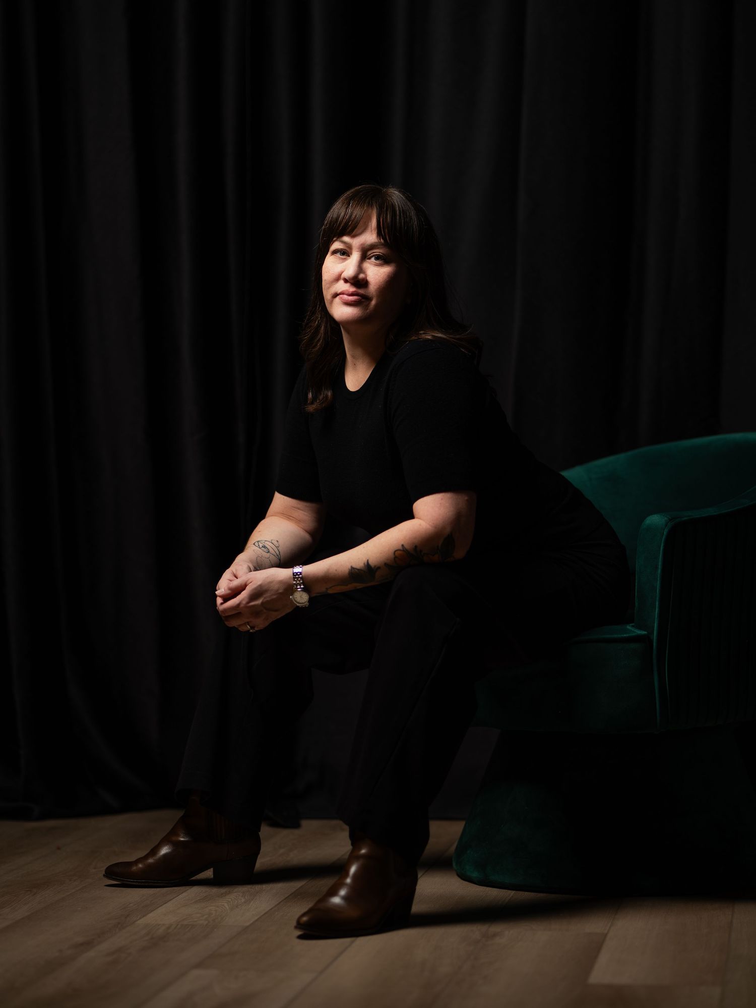 A person in all black attire sits on a wooden floor against a dark backdrop with an emerald green velvet chair nearby.