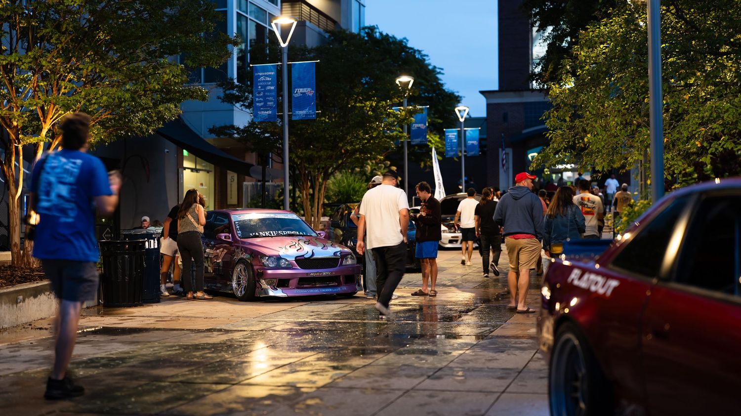 Cars parked along illuminated city street at dusk with pedestrians walking by.