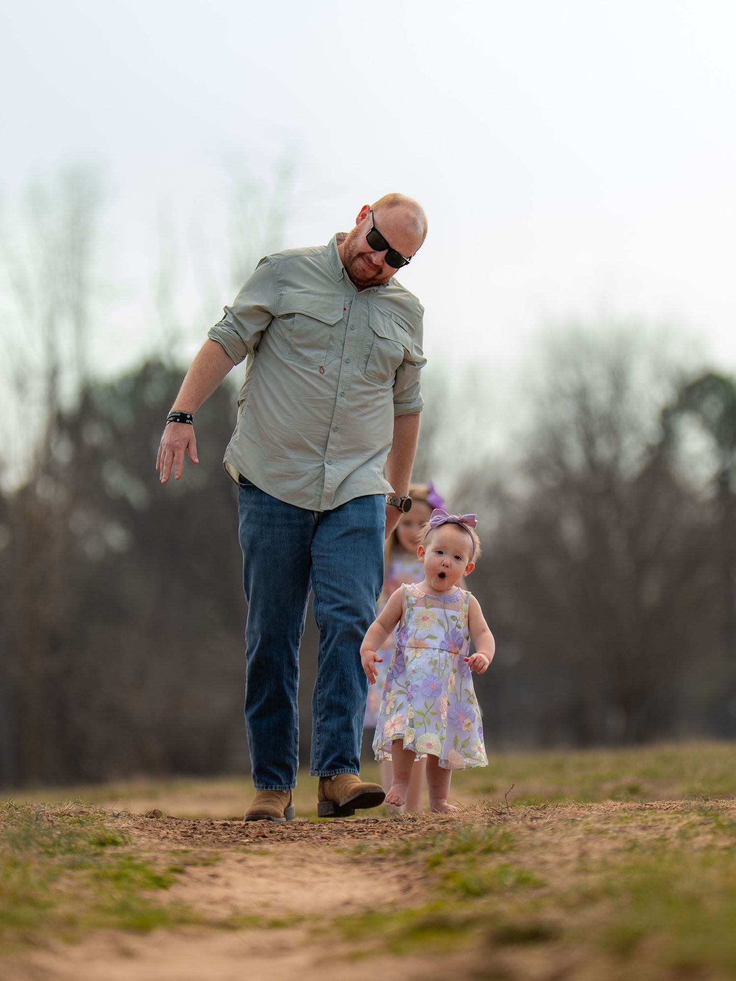A man walks with two young girls on a dirt path outdoors, with bare trees in the background.