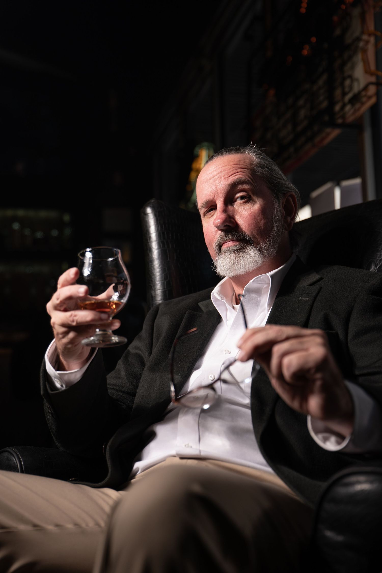 Sophisticated older man in black blazer holding whiskey glass and cigar while relaxing in leather chair in dark setting.